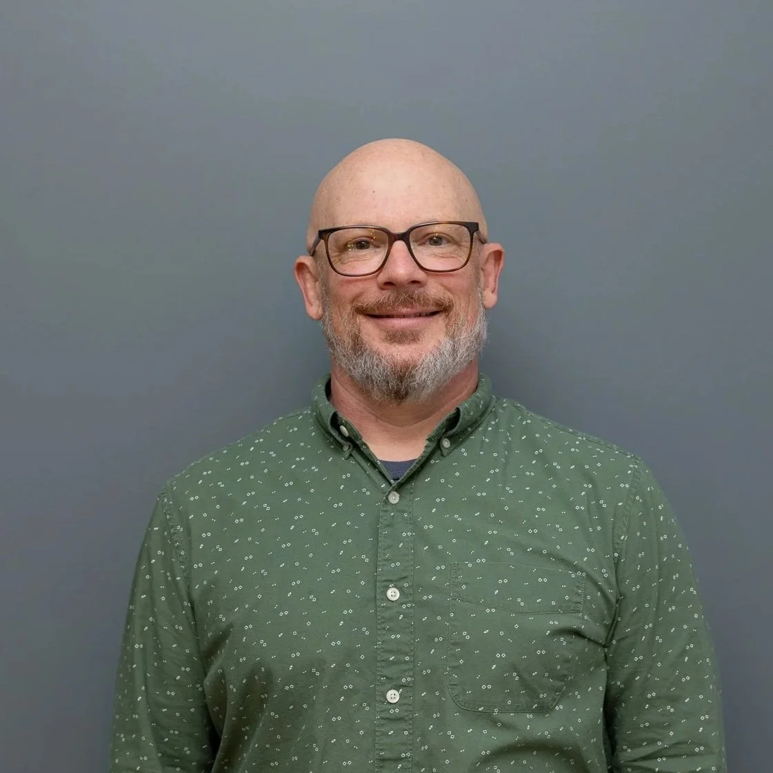 A smiling, bald man with glasses and a gray beard, wearing a green button-up shirt with a small white pattern, standing against a plain gray background.