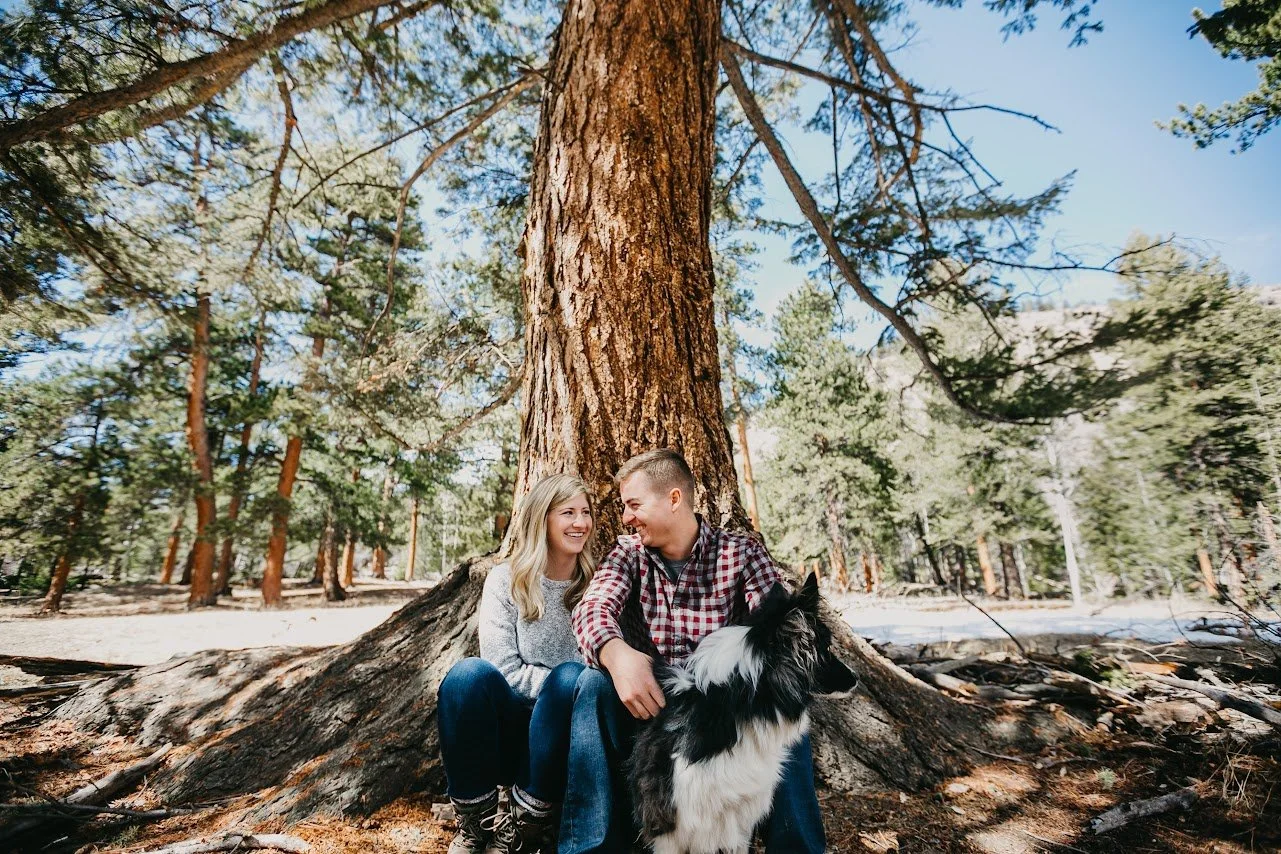 A smiling couple sitting on the ground in front of a large tree in a wooded area, with their black and white dog, enjoying a sunny day outdoors.