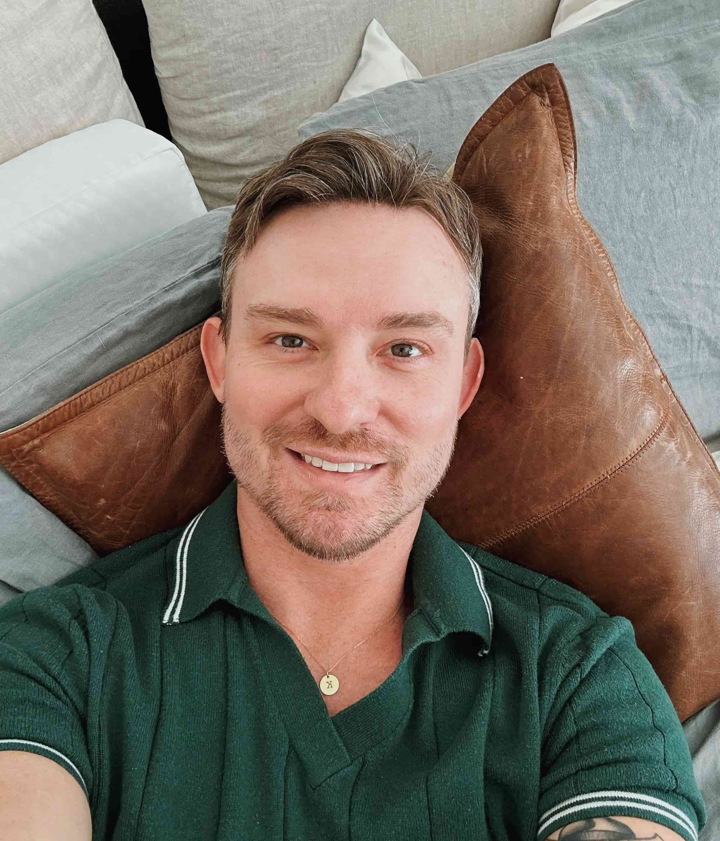 A man with short hair and a green shirt smiles on a bed with gray and brown pillows, feeling supported by LGBT Therapy. Kris Thorpe LCSW