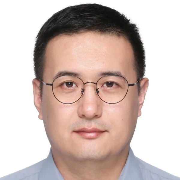 Headshot of a man with short black hair, wearing round glasses and a light blue shirt, looking directly at the camera against a white background.