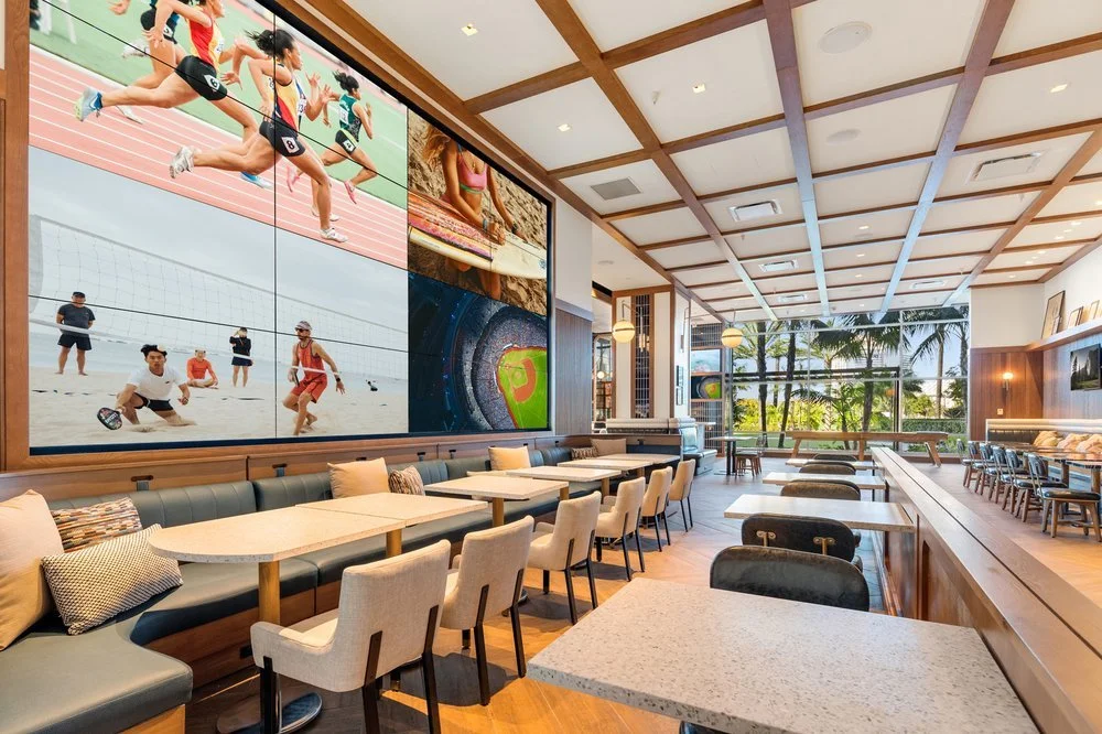 Modern lounge with large wall-mounted sports screen, tables and chairs, and windows showing palm trees outside.