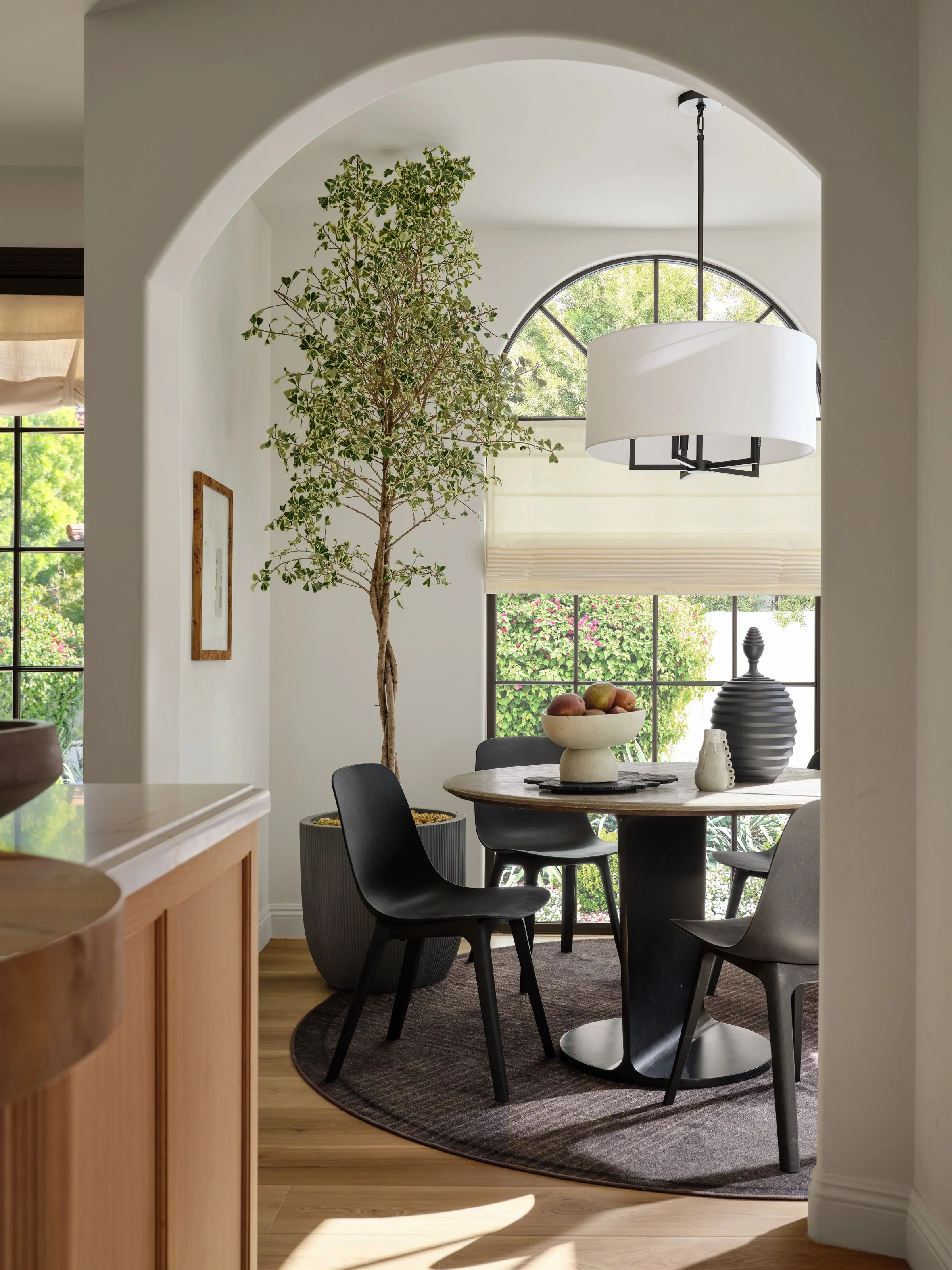 A light-filled dining space that balances architectural character with understated elegance. Soft curves, warm wood tones, sculptural furnishings, and bold black accents create a refined yet inviting setting, while the oversized greenery brings an or