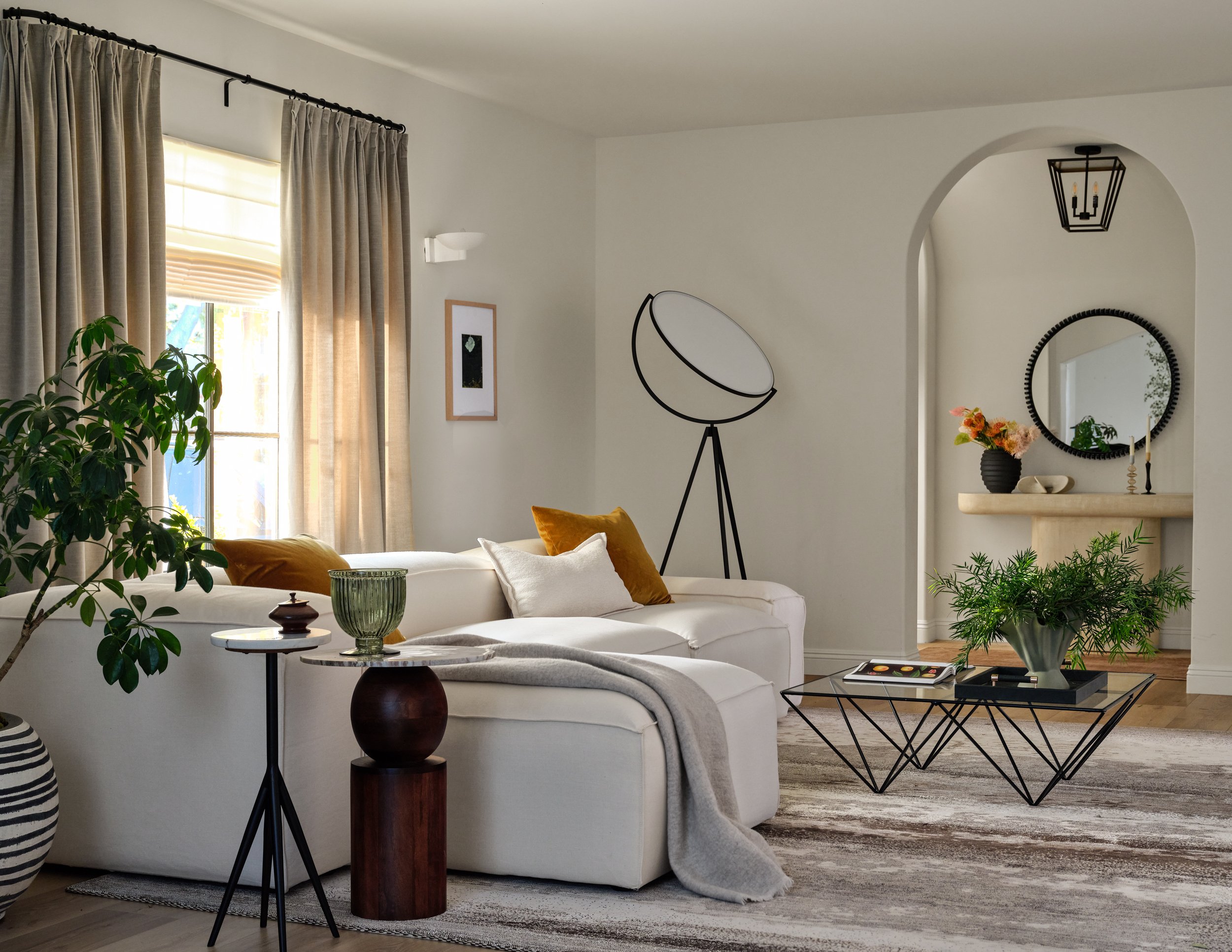 A warm, light-filled living room that balances comfort with sculptural design. Soft neutrals, rich wood tones, organic greenery, and touches of ochre create a layered, inviting space, while the arched opening and statement lighting add architectural 