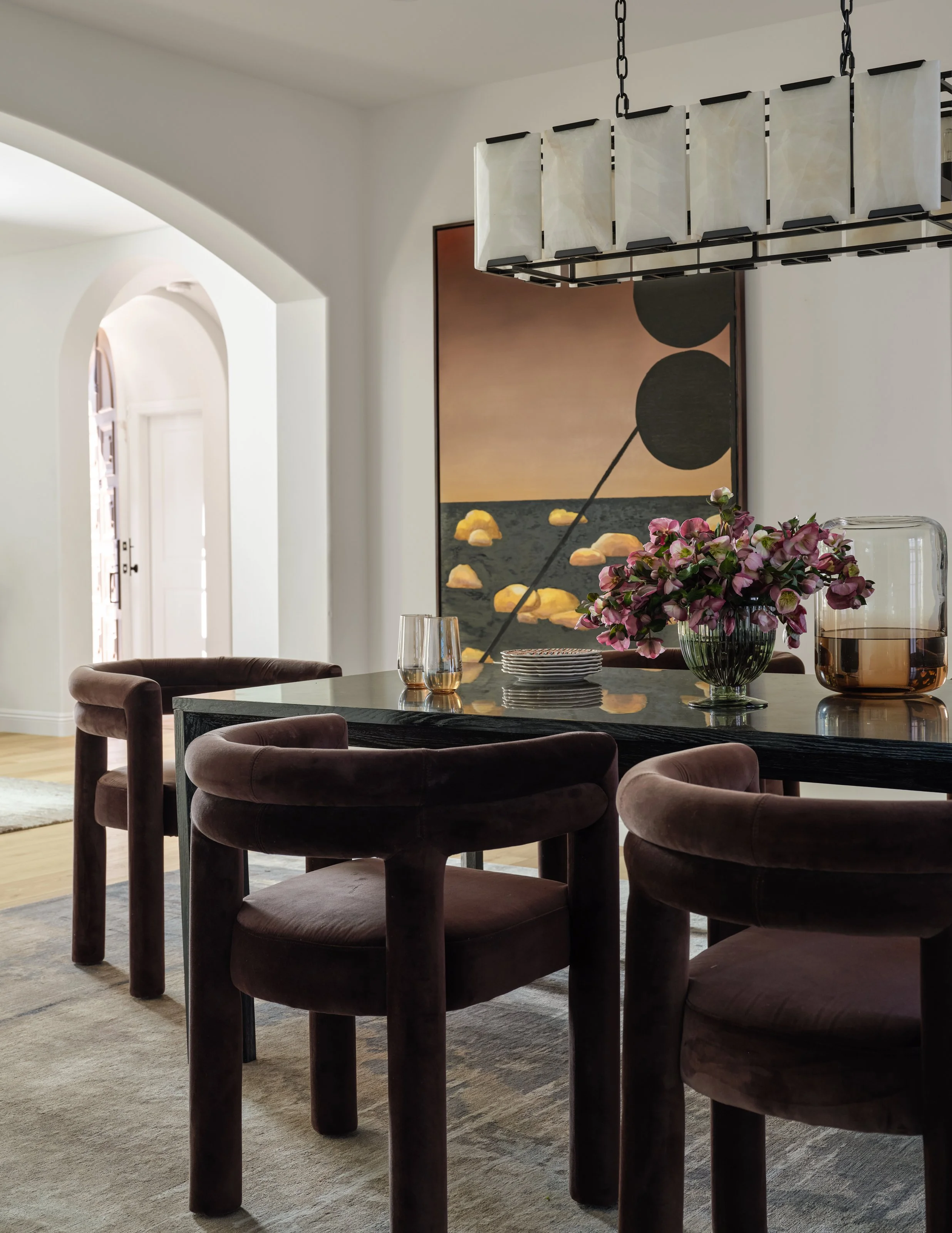 A dramatic dining space grounded in rich chocolate velvet, sculptural lighting, and expressive oversized art. The deeper tones bring warmth and intimacy, while the soft arches and natural light keep the room feeling open, refined, and effortlessly in