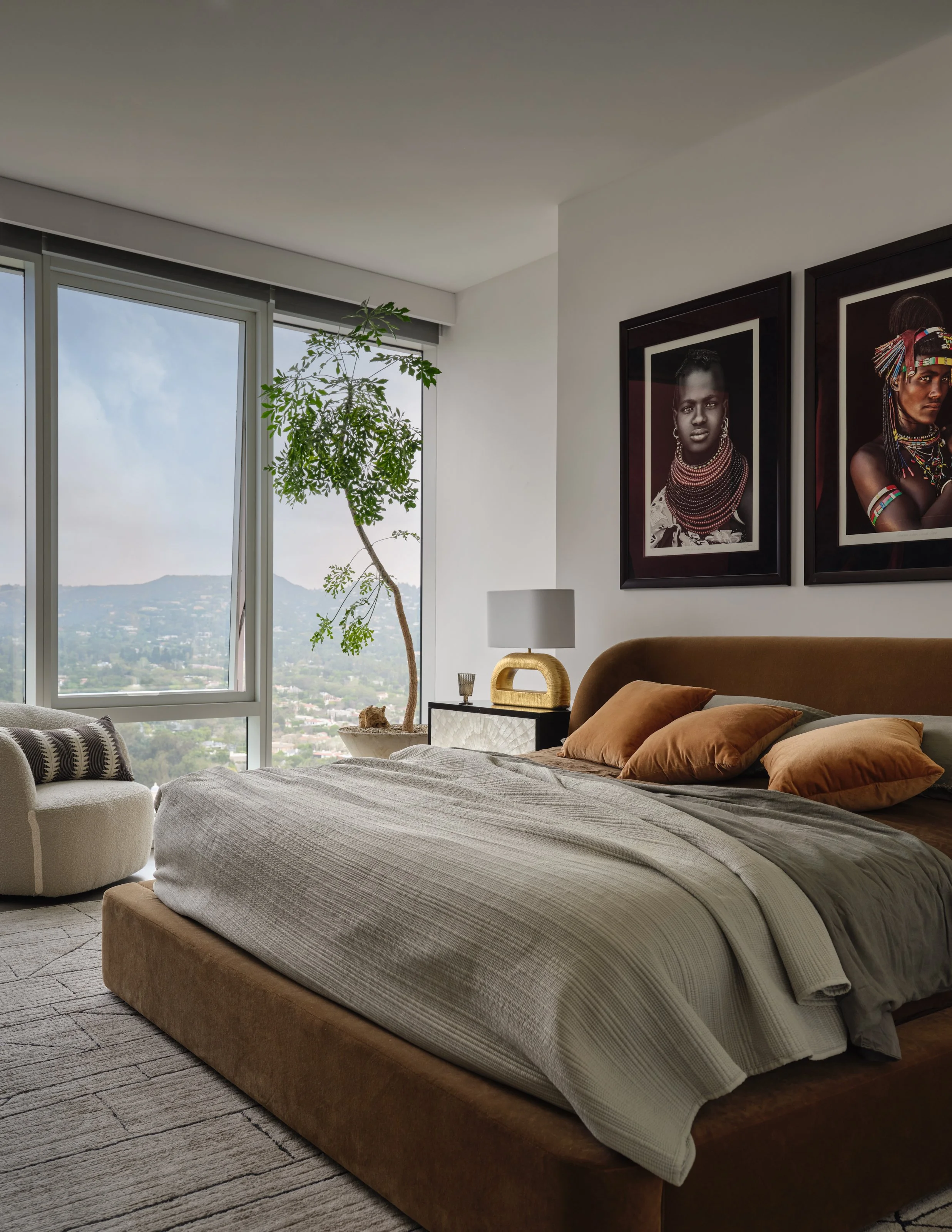 A serene primary bedroom grounded in warm, earthy tones and soft layers of texture. The upholstered bed creates a sense of comfort, while expressive portraiture, sculptural furnishings, and organic greenery bring culture and personality to the space.