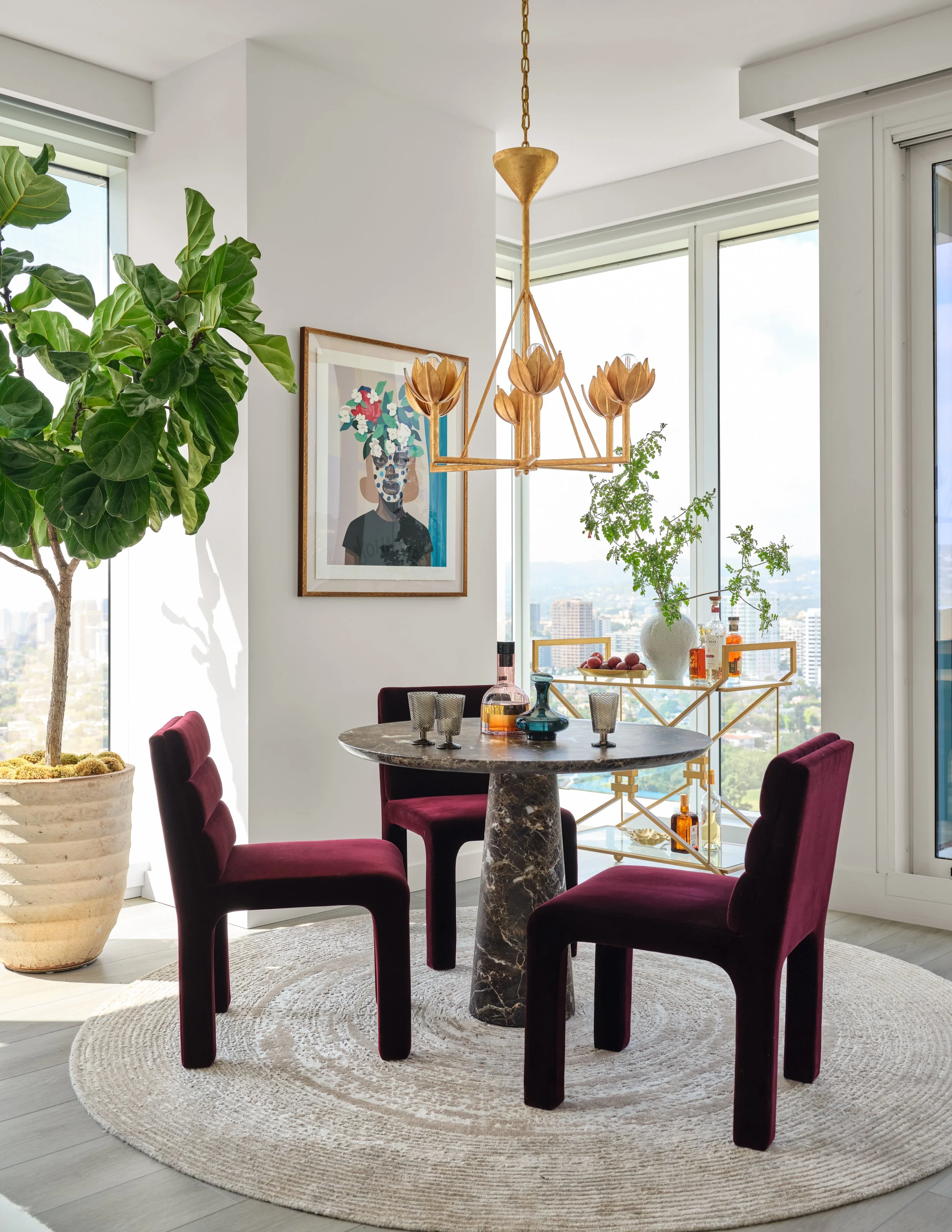 An intimate dining moment framed by expansive city views. Rich burgundy velvet chairs and a sculptural marble table bring warmth and depth to the light-filled space, while the statement brass chandelier, curated art, and elegant bar cart add a sense 