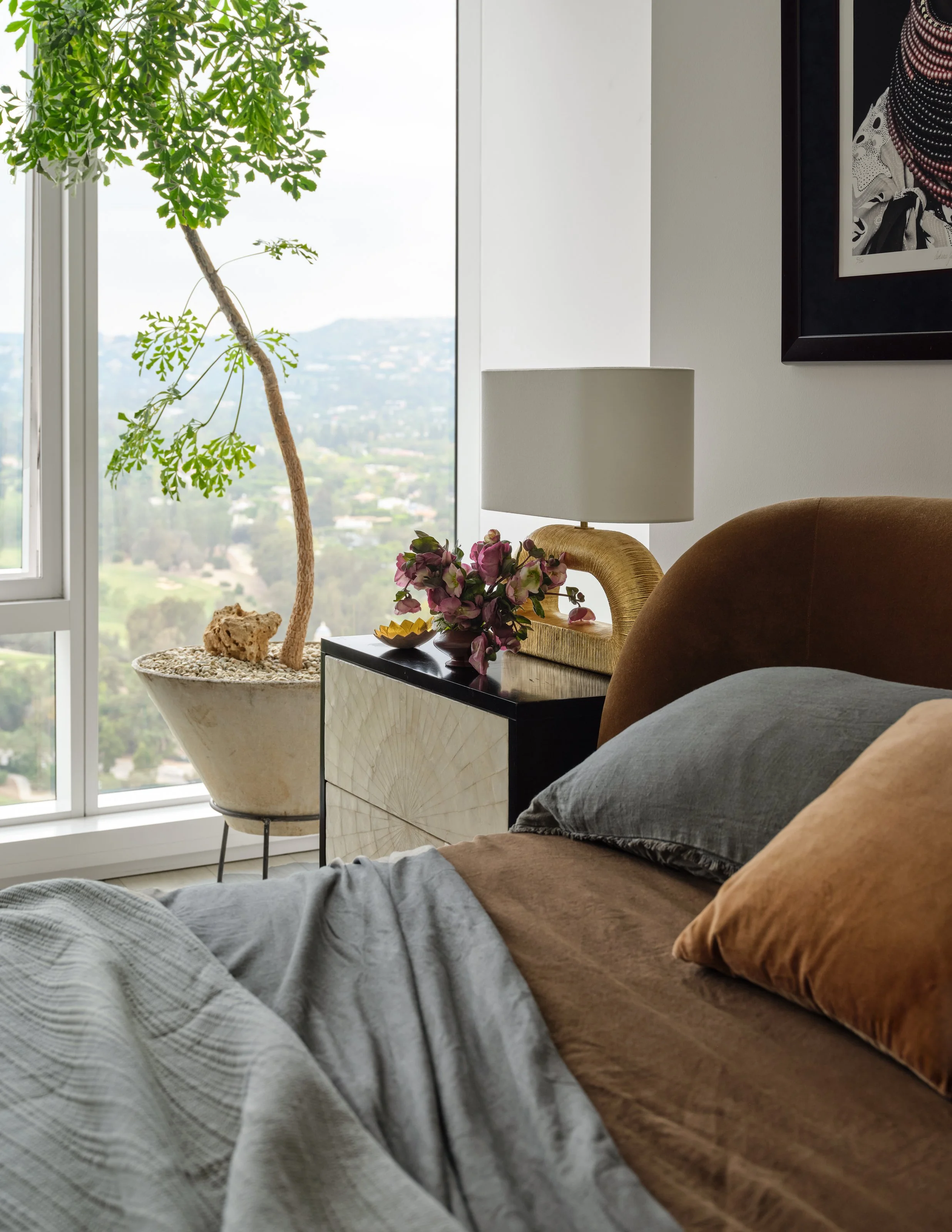 A closer look at the primary bedroom highlights the layered materials and thoughtful details that give the space its warmth. Rich caramel upholstery, soft gray linens, a sculptural bedside lamp, and natural greenery create an intimate moment against 