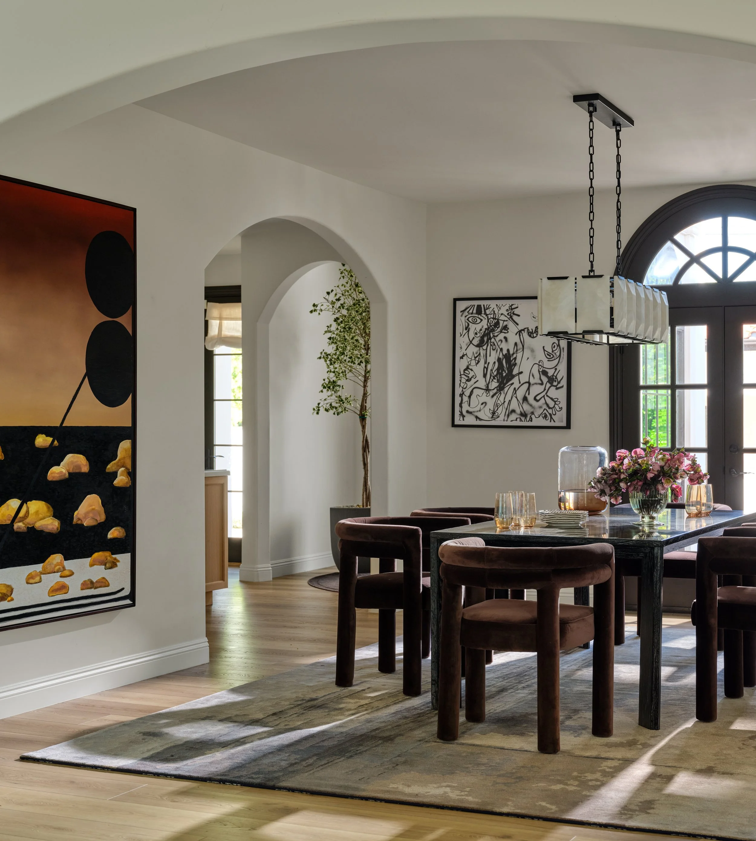 A sophisticated dining space where bold art, sculptural forms, and rich chocolate tones create a sense of warmth and drama. Soft architectural arches and natural light balance the darker furnishings, giving the room an elevated yet inviting feel.
