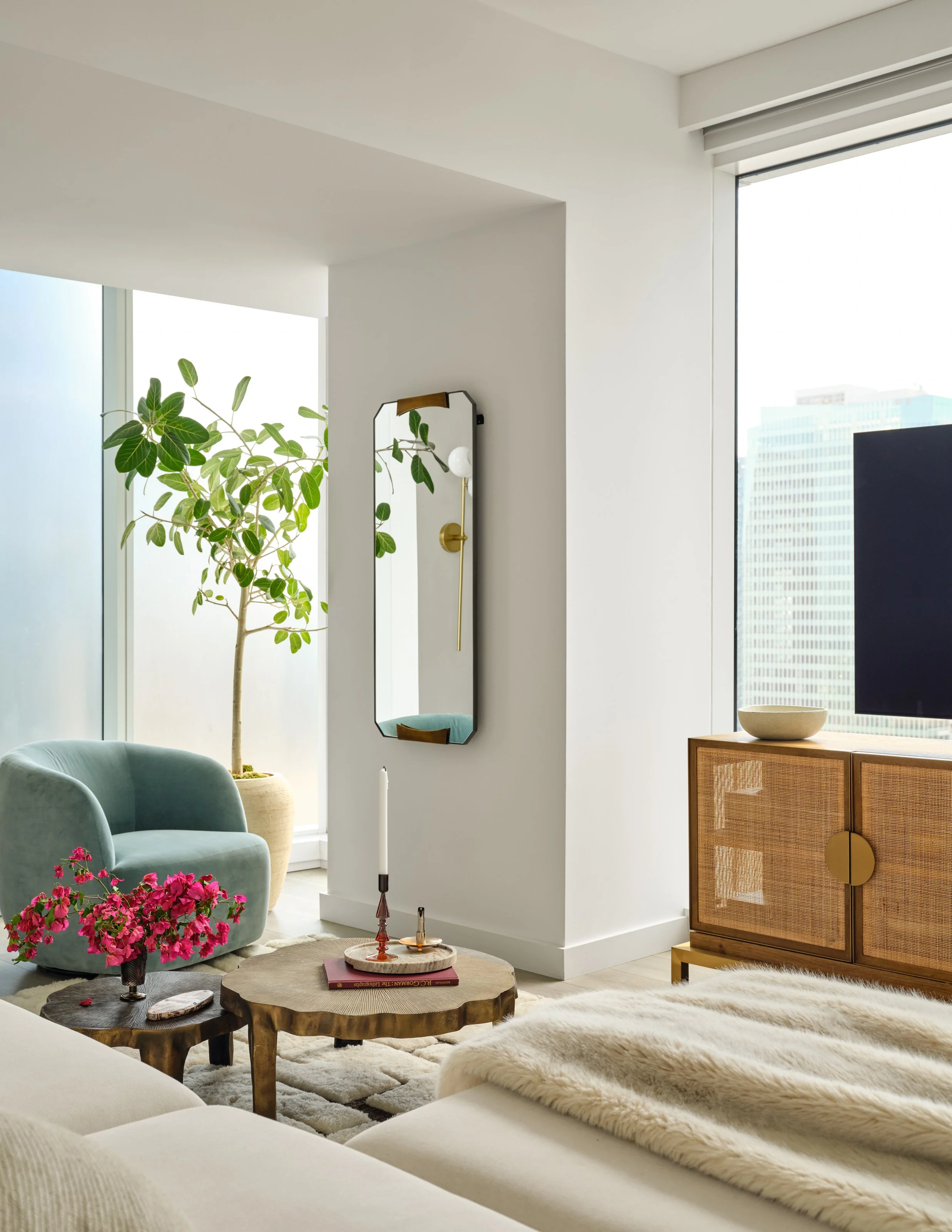 A light-filled living space that balances softness with sculptural detail. The curved blue velvet chair, woven media console, warm brass accents, and layered textures bring personality and warmth to the neutral palette, while greenery and vibrant flo