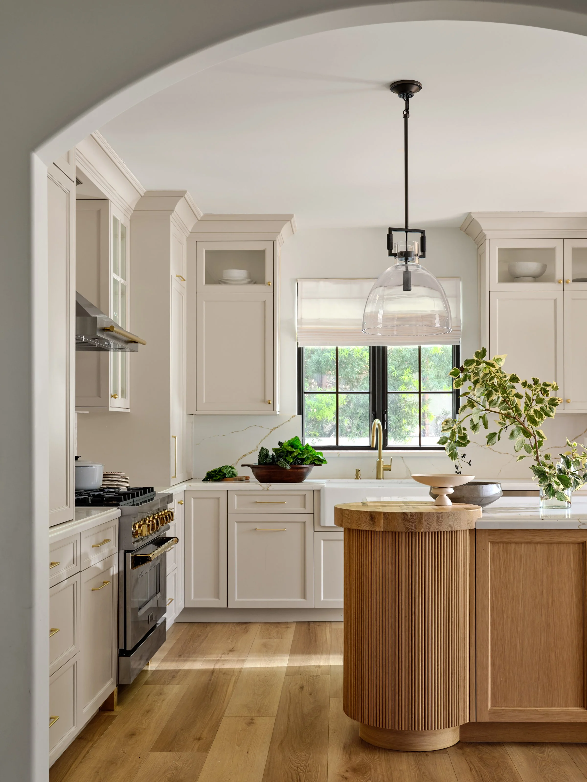 A warm, refined kitchen where classic details meet modern craftsmanship. Soft cabinetry, natural oak, fluted wood detailing, and brushed brass accents create a layered, timeless feel, while the sculptural island and arched entry bring softness and ar