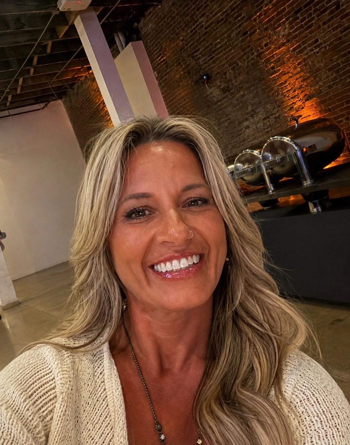 A smiling woman with shoulder-length blond hair wearing a white sleeveless top, standing against a gray textured wall.
