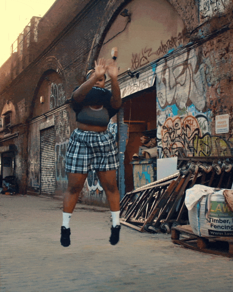 A person jumping in the air with arms raised, in an urban alleyway with graffiti-covered walls, metal barriers, and construction materials.