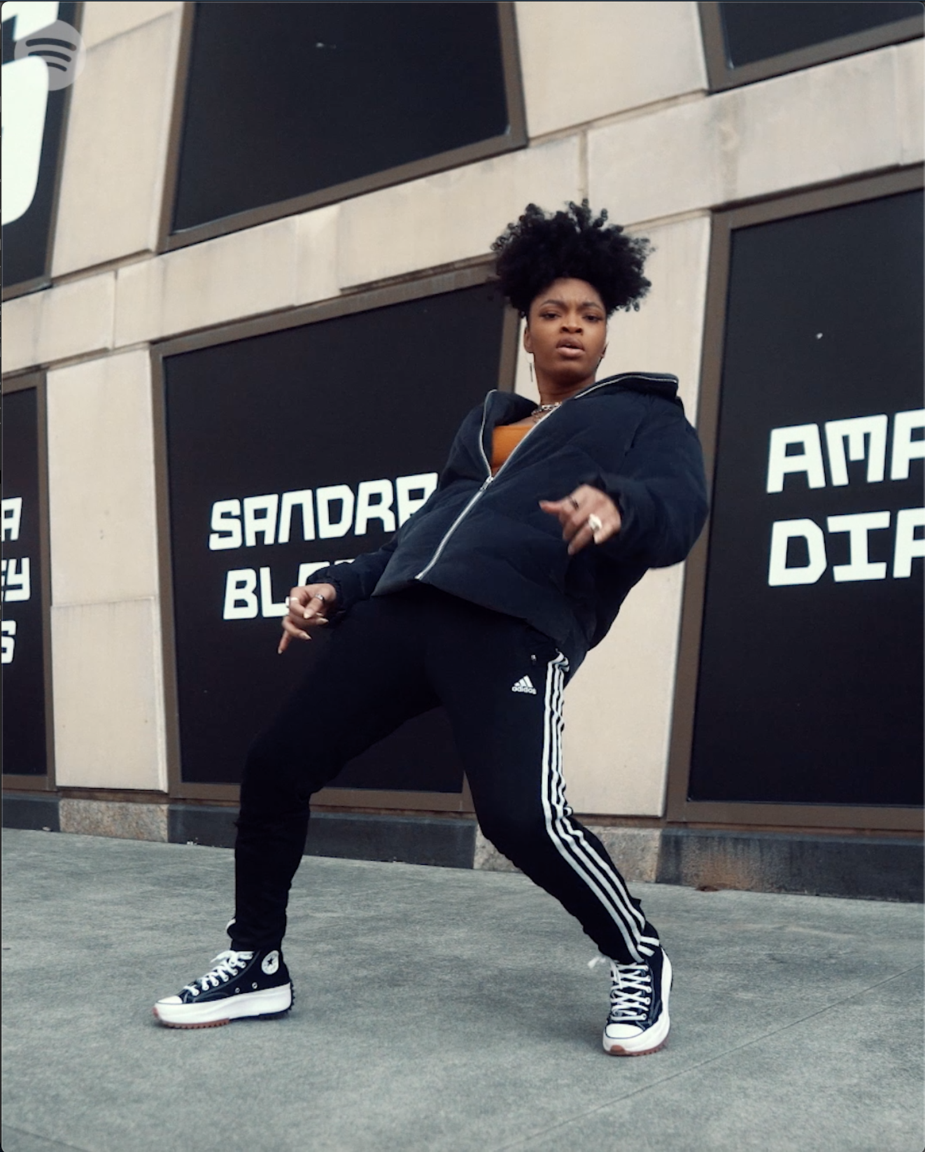 A woman with curly hair wearing a black jacket, black Adidas sweatpants, and black and white Converse sneakers, dancing outside a building with large windows and signs that read 'Sandra' and 'Black' for Spotify's African Heat campaign launch.