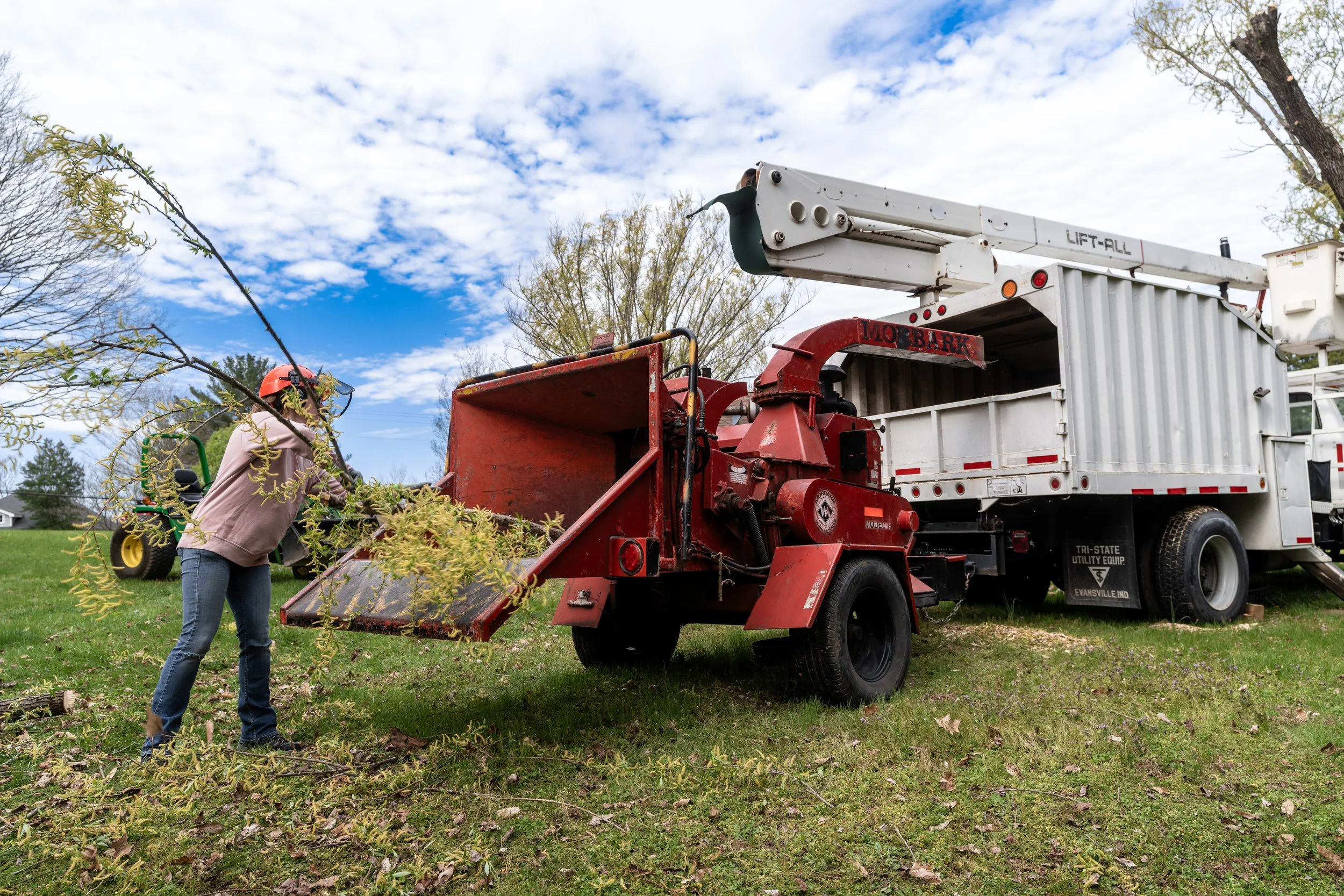 A person wearing a pink jacket, jeans, and a safety helmet with face shield operating a wood chipper in a grassy yard, with a tree branch being fed into the machine, and a truck with a hydraulic lift nearby under a partly cloudy sky.
