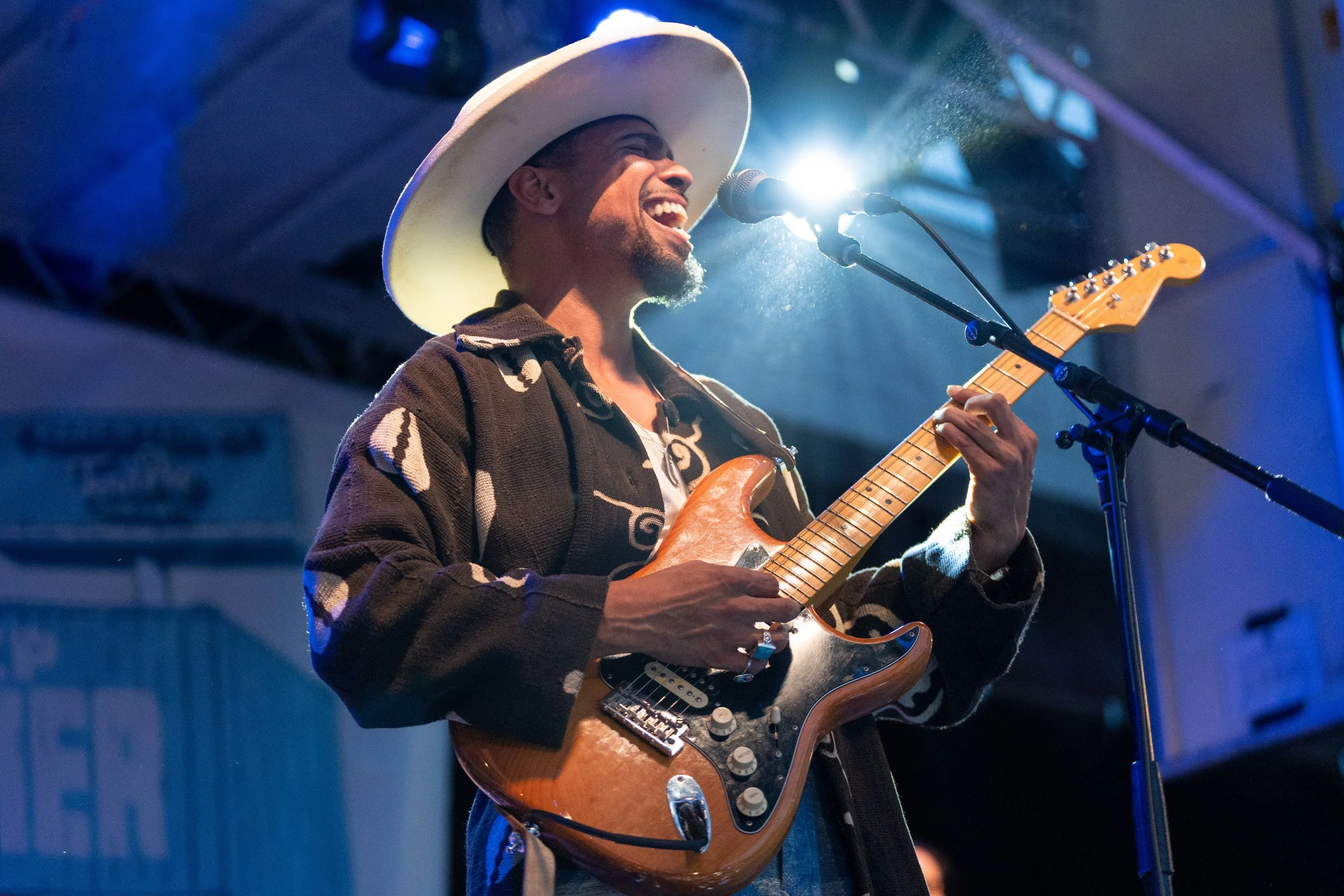 A musician wearing a wide-brimmed hat and a patterned jacket, singing passionately into a microphone while playing an electric guitar on stage, with blue stage lighting.