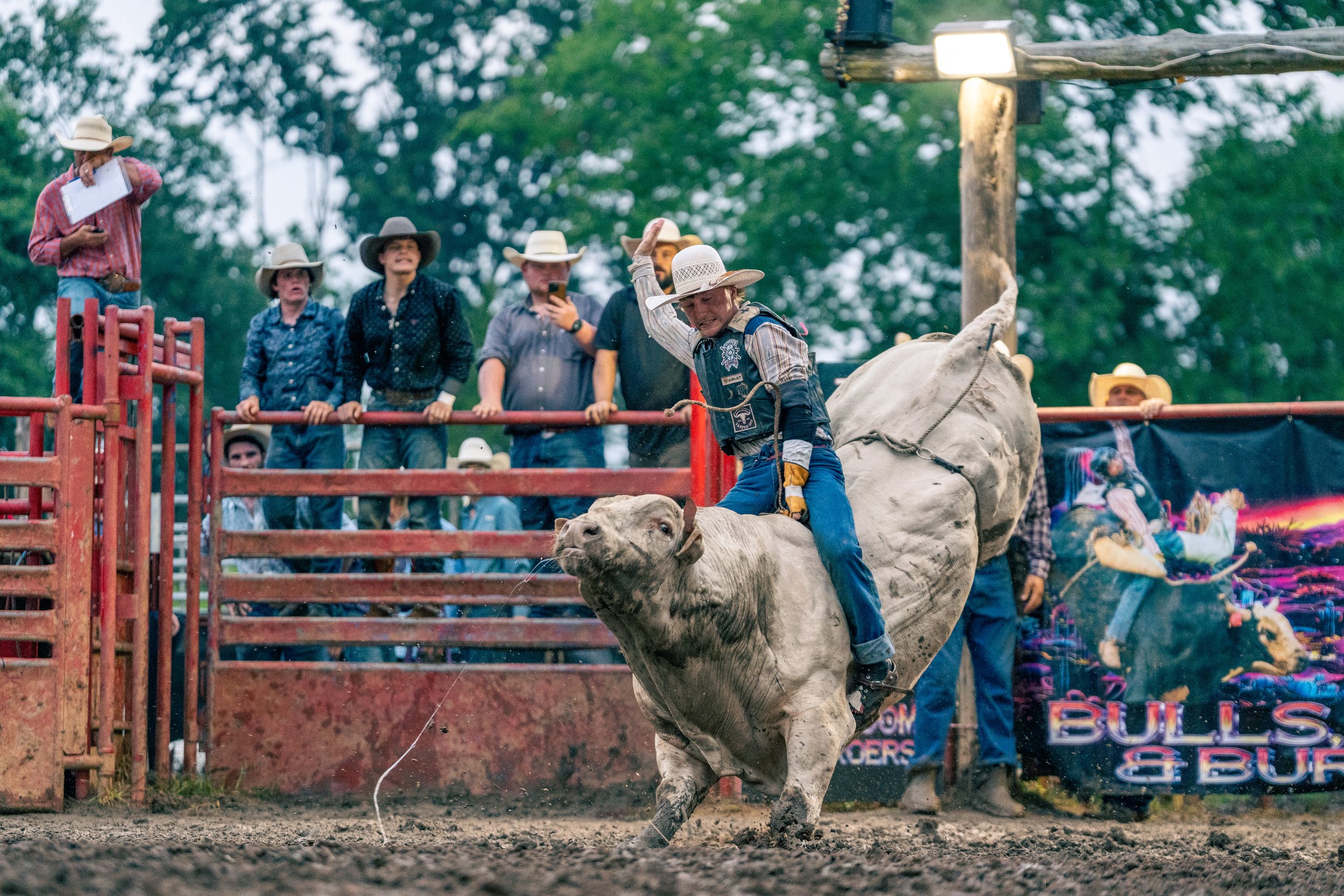 A rodeo scene with a bull rider holding on to a bull during a rodeo event, surrounded by onlookers wearing cowboy hats.