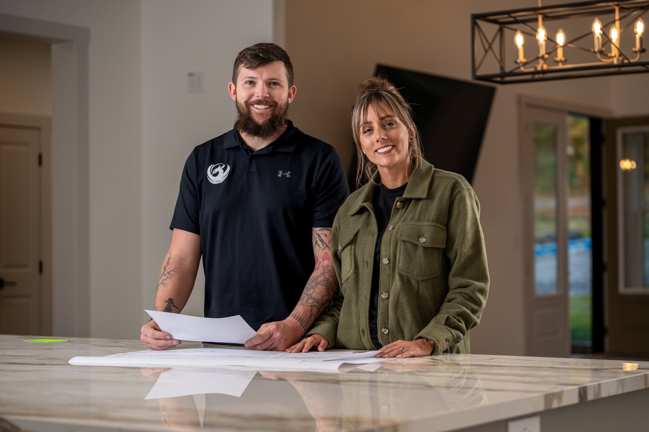 A man and woman with tattoos on their arms are smiling at the camera while reviewing documents on a table in a well-lit room with large windows and a black chandelier.