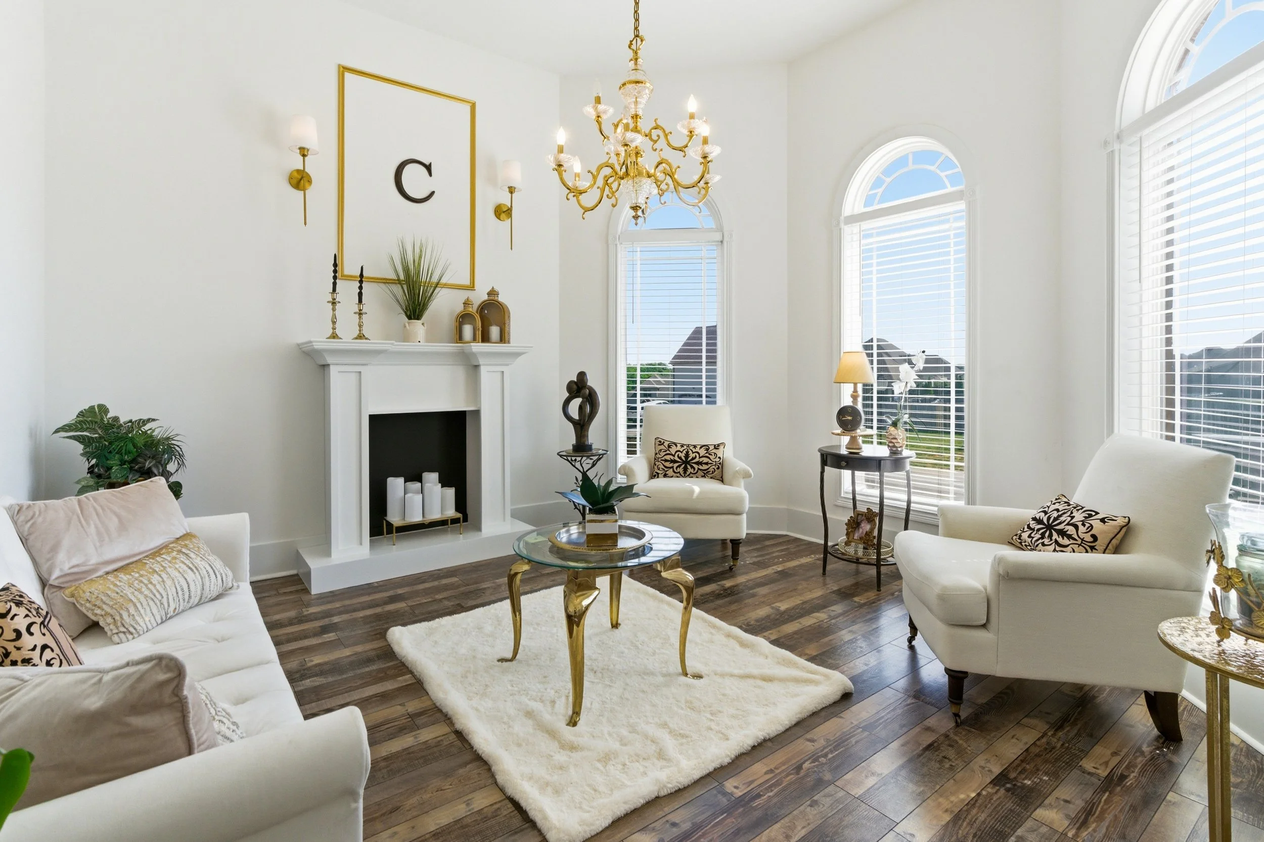 A bright living room with white walls and large arched windows. It features a gold chandelier, a white fireplace, and elegant furniture including white sofas and armchairs with decorative pillows. There are gold accents, potted plants, candles, and a rug on a dark wood floor.