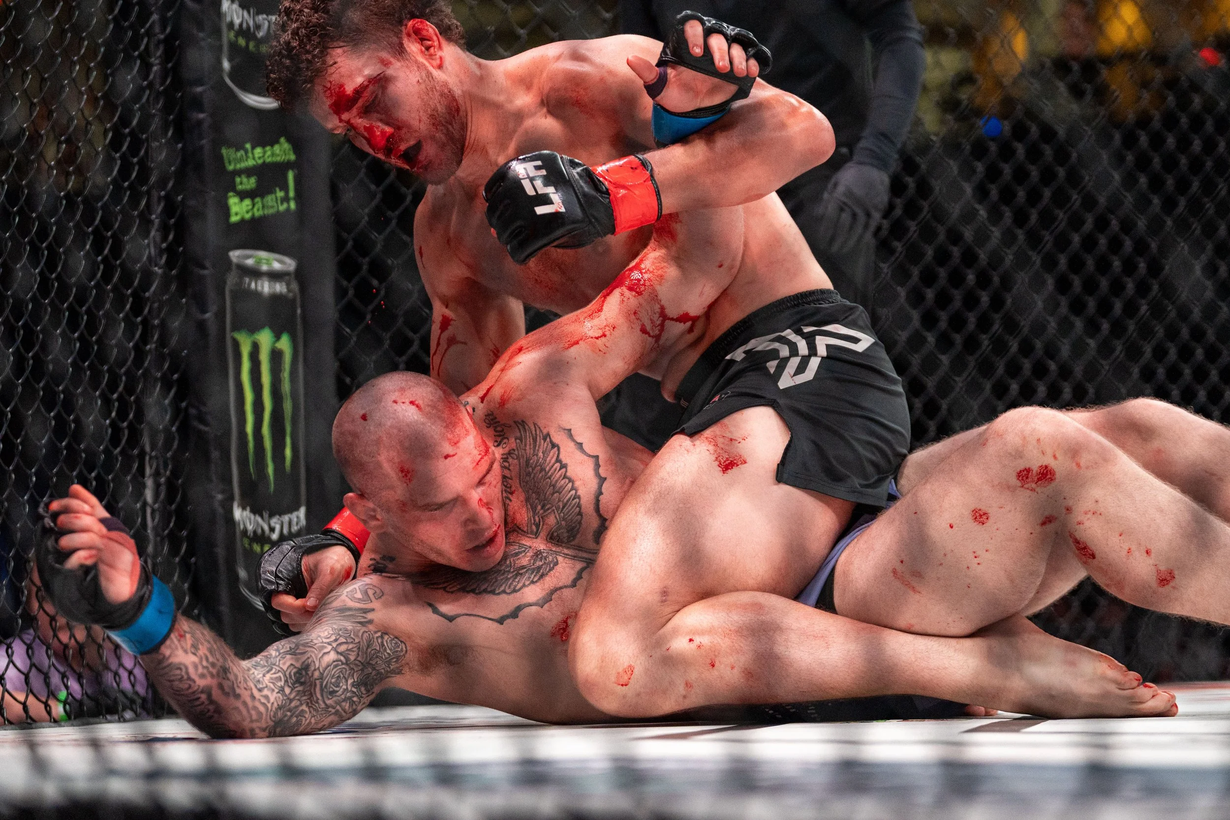 Two mixed martial arts fighters engaged in a bloody fight inside an octagon cage, with one fighter on top and the other on the ground, surrounded by a black mesh fence.