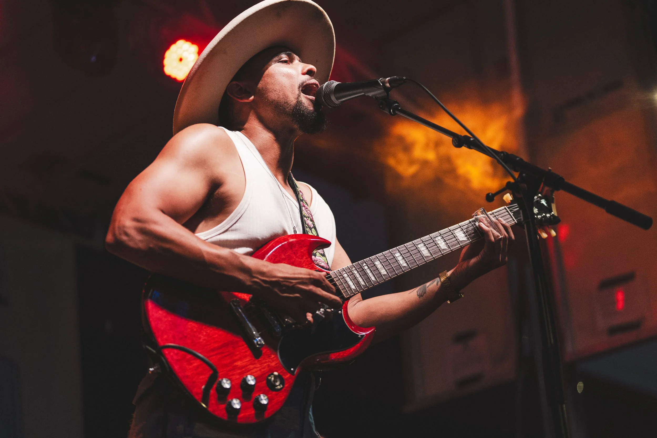 A man wearing a wide-brimmed hat and a sleeveless shirt, singing into a microphone while playing a red electric guitar on stage, with warm stage lighting in the background.