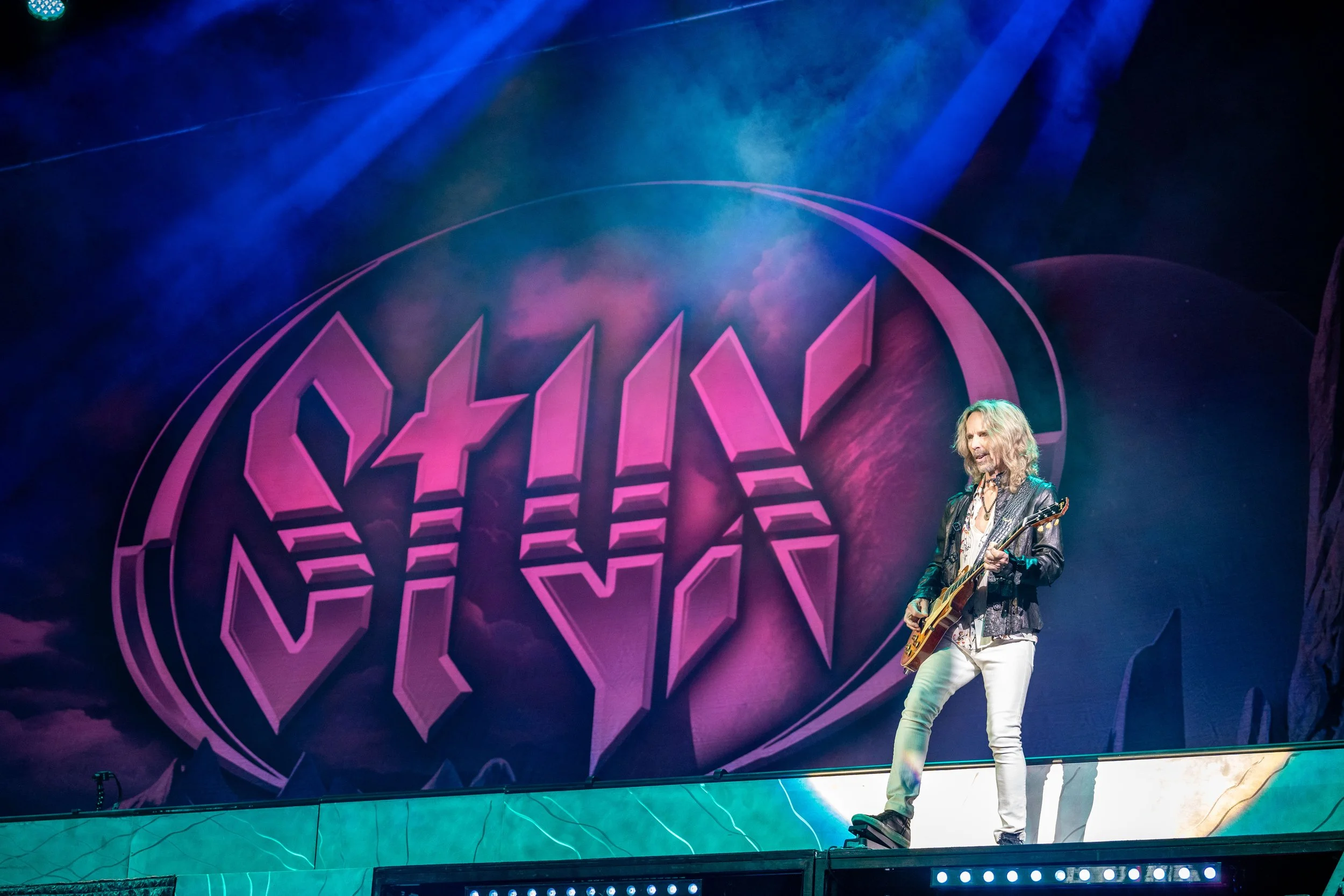 A musician with long blonde hair playing a guitar on stage with a neon pink and purple background.