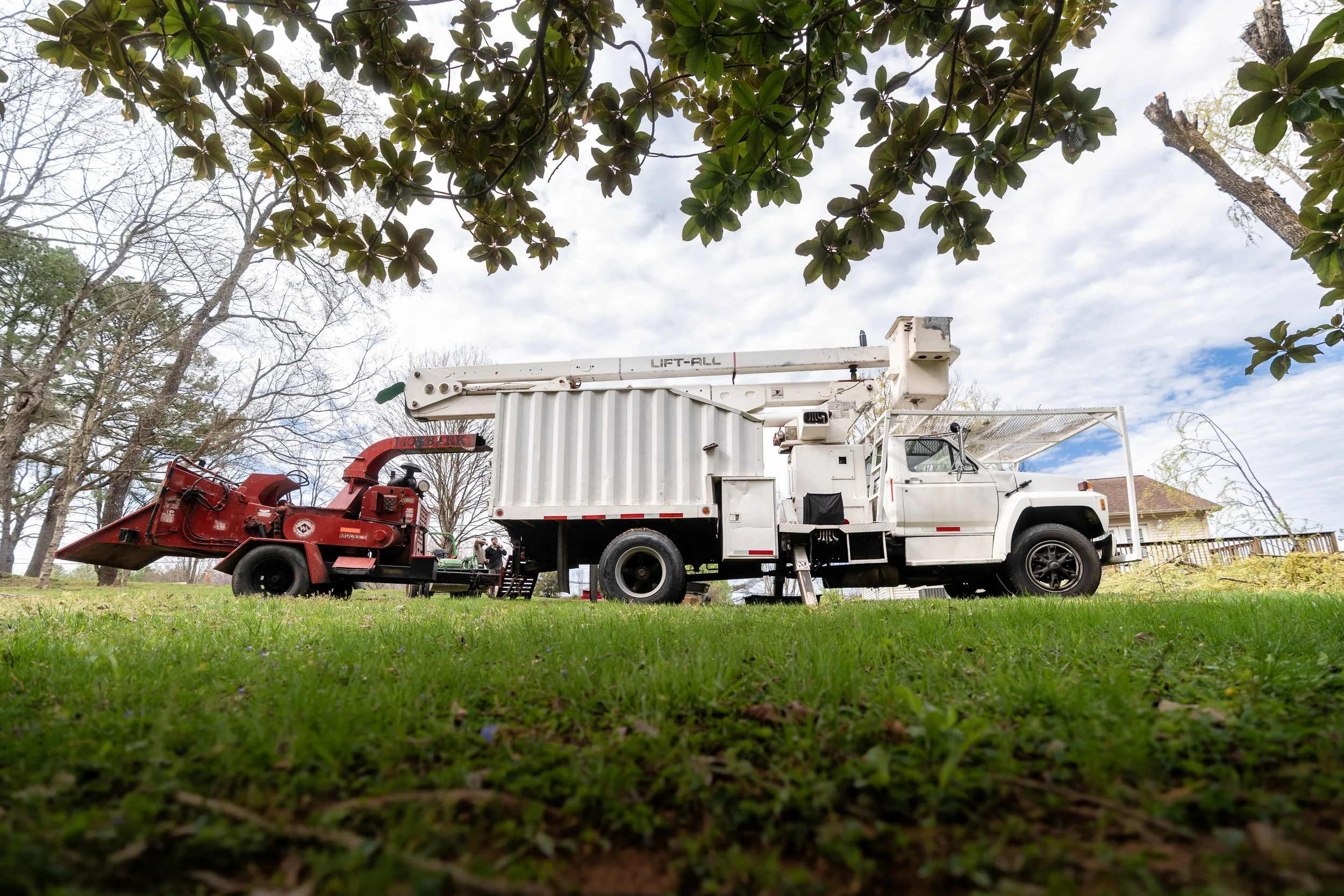 A white utility truck with an extended crane arm parked on grass under a large leafy tree, with a cloudy sky in the background.