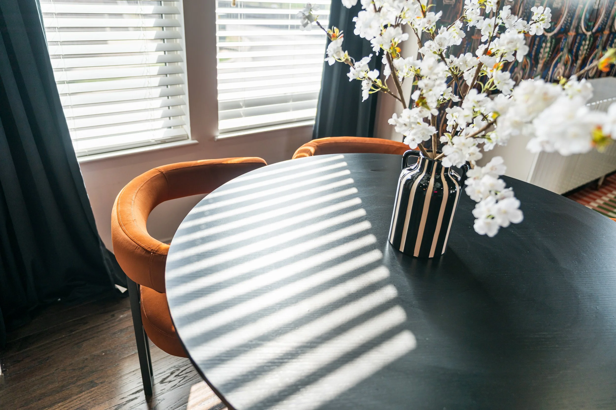 A dining room table with a striped black and white vase holding white flowers. Sunlight streams through window blinds casting shadows on the table. There are two brown chairs around the table and curtains in the background.