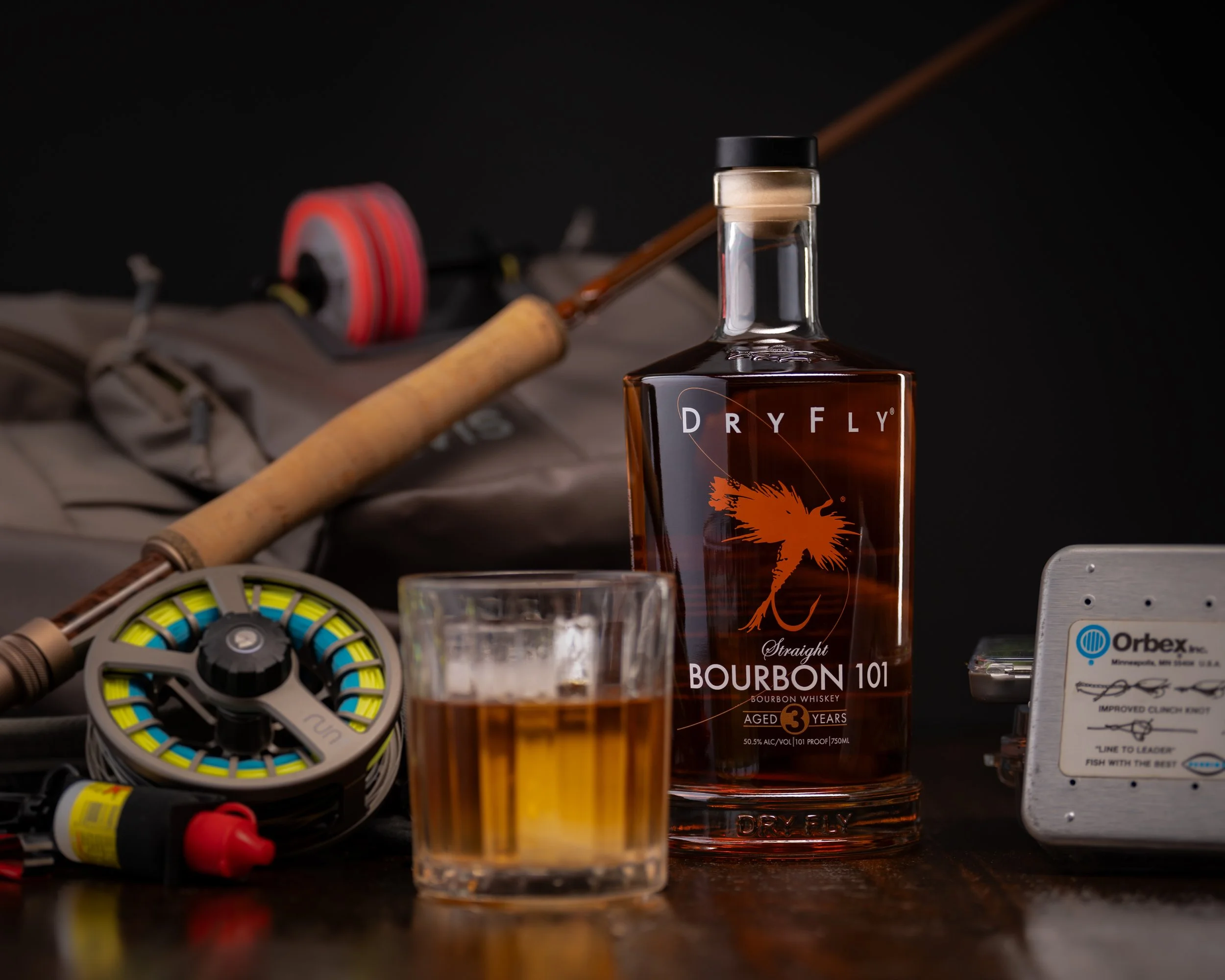 A bottle of Dry Fly bourbon whiskey with a glass of whiskey in front, surrounded by fishing equipment including reels and a line spool, against a dark background.