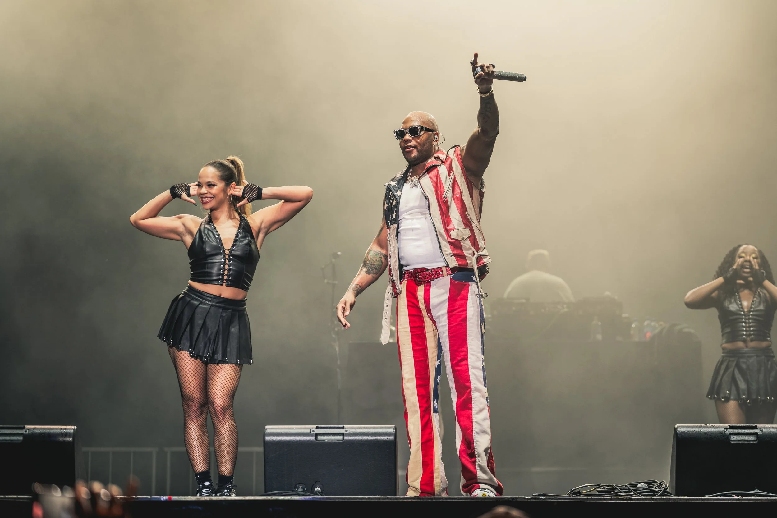 A male performer wearing sunglasses and an American flag-themed outfit holding a microphone on stage, flanked by two female performers dressed in black metallic outfits, with other performers faintly visible in the background.