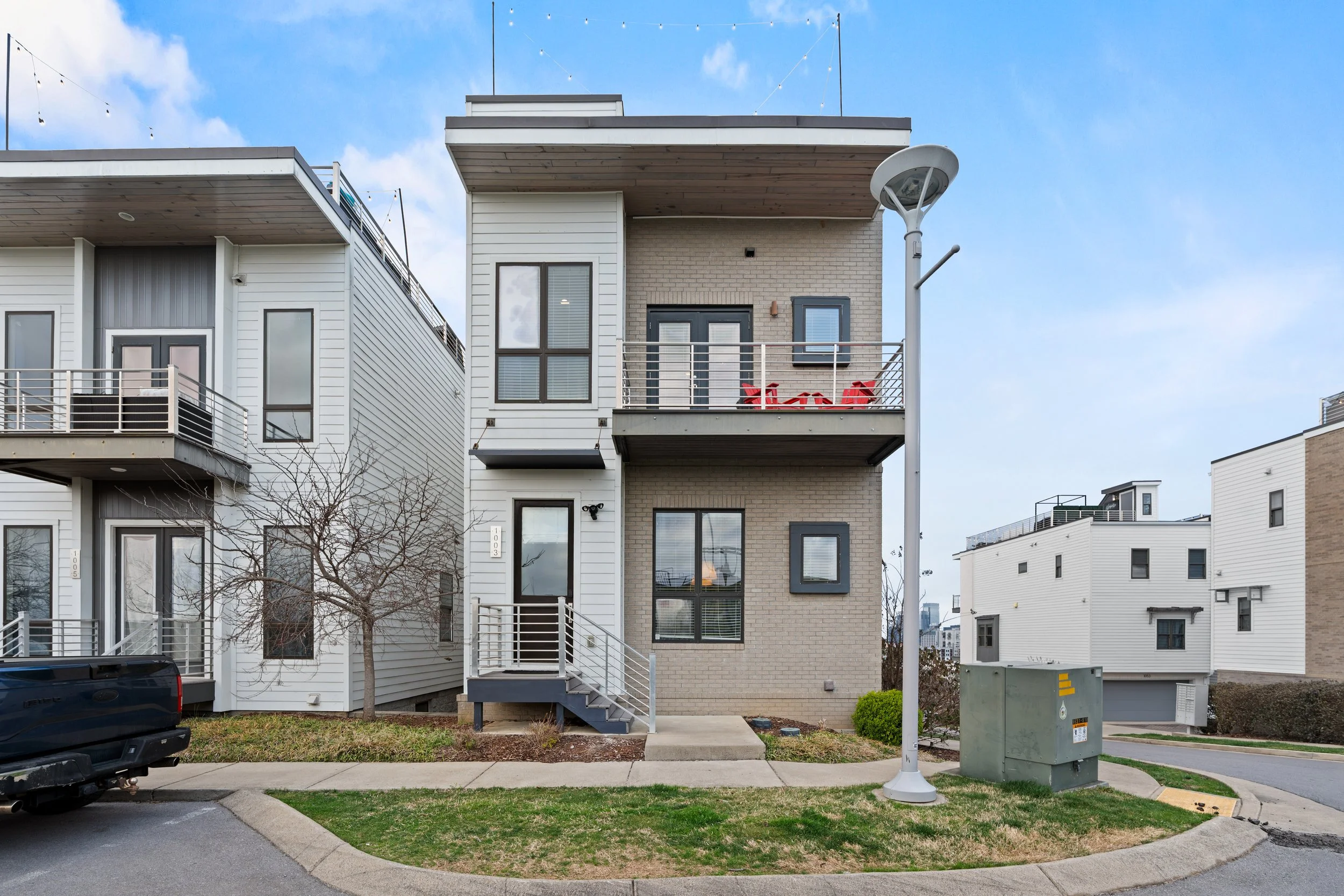Modern multi-story residential building with white and beige exterior, multiple balconies with red chairs, large windows, and a tree in front. There is a streetlamp, a utility box, and a parked vehicle. The sky is partly cloudy.