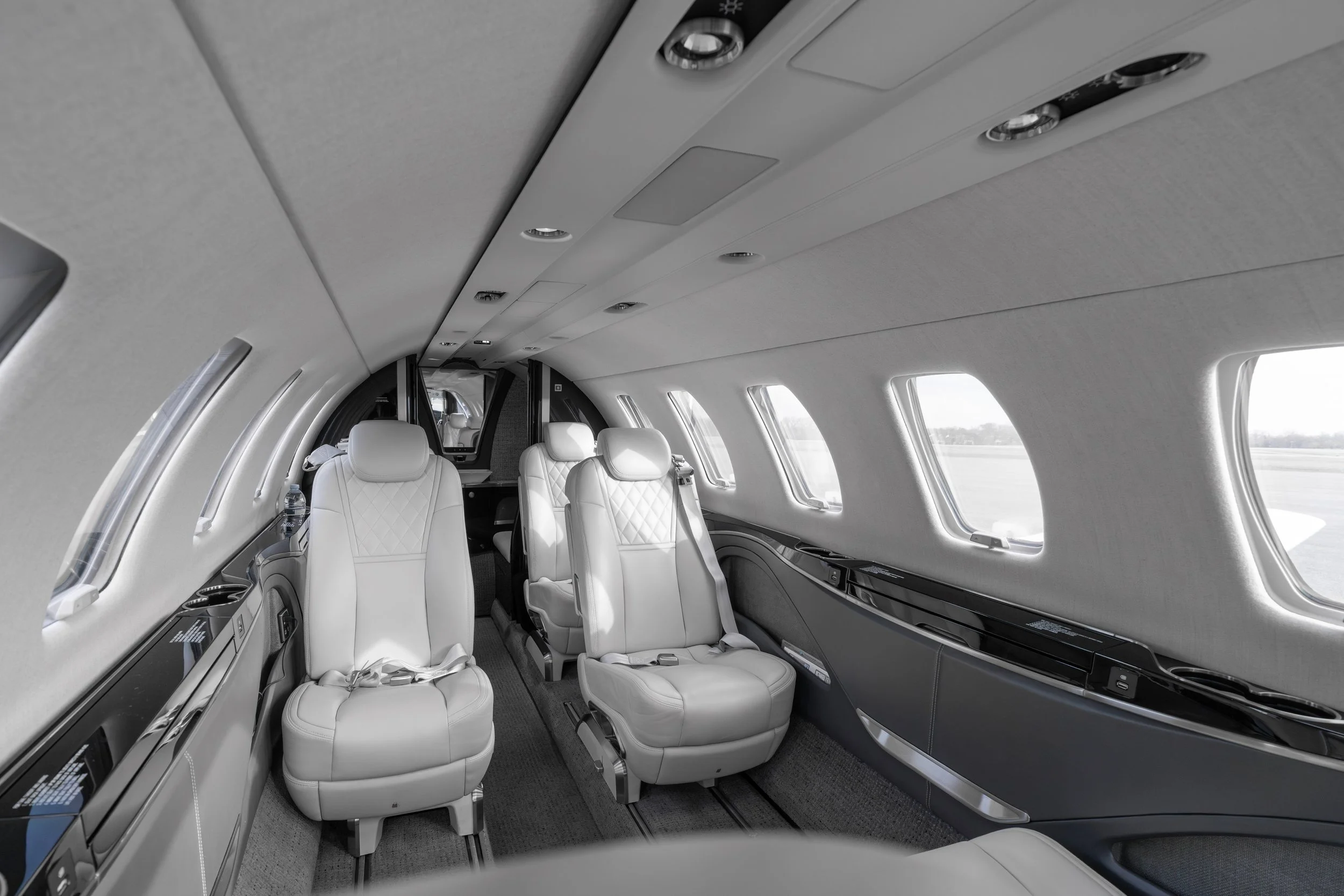 Interior of a private jet with white leather seats, overhead lighting, windows along the side, and a streamlined ceiling with control panels.