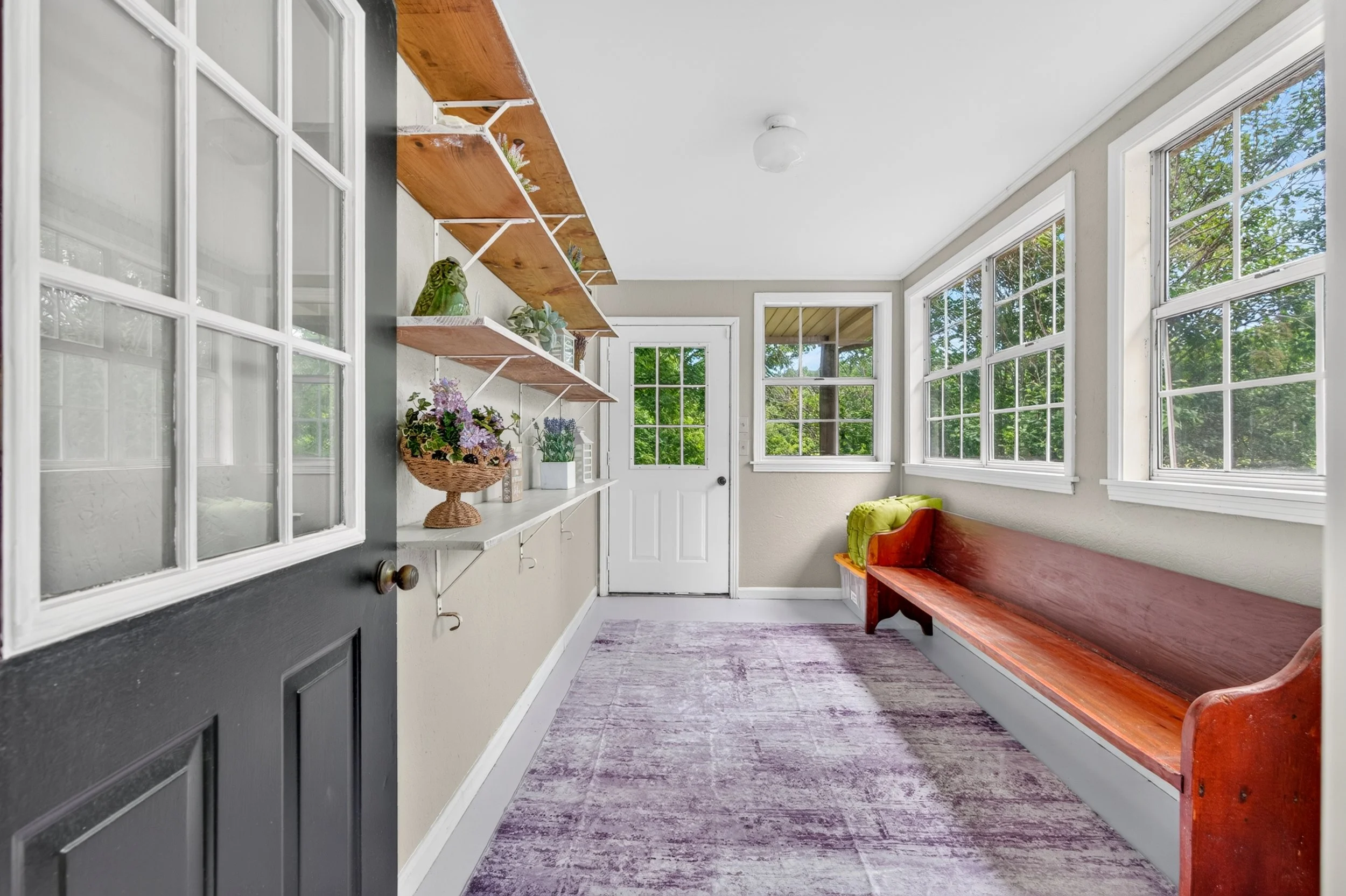 A bright sunroom with large windows showing green trees outside, wooden built-in bench with a green pillow, floating shelves with plants and decorative items, a pink and purple rug, and a light fixture on the ceiling.