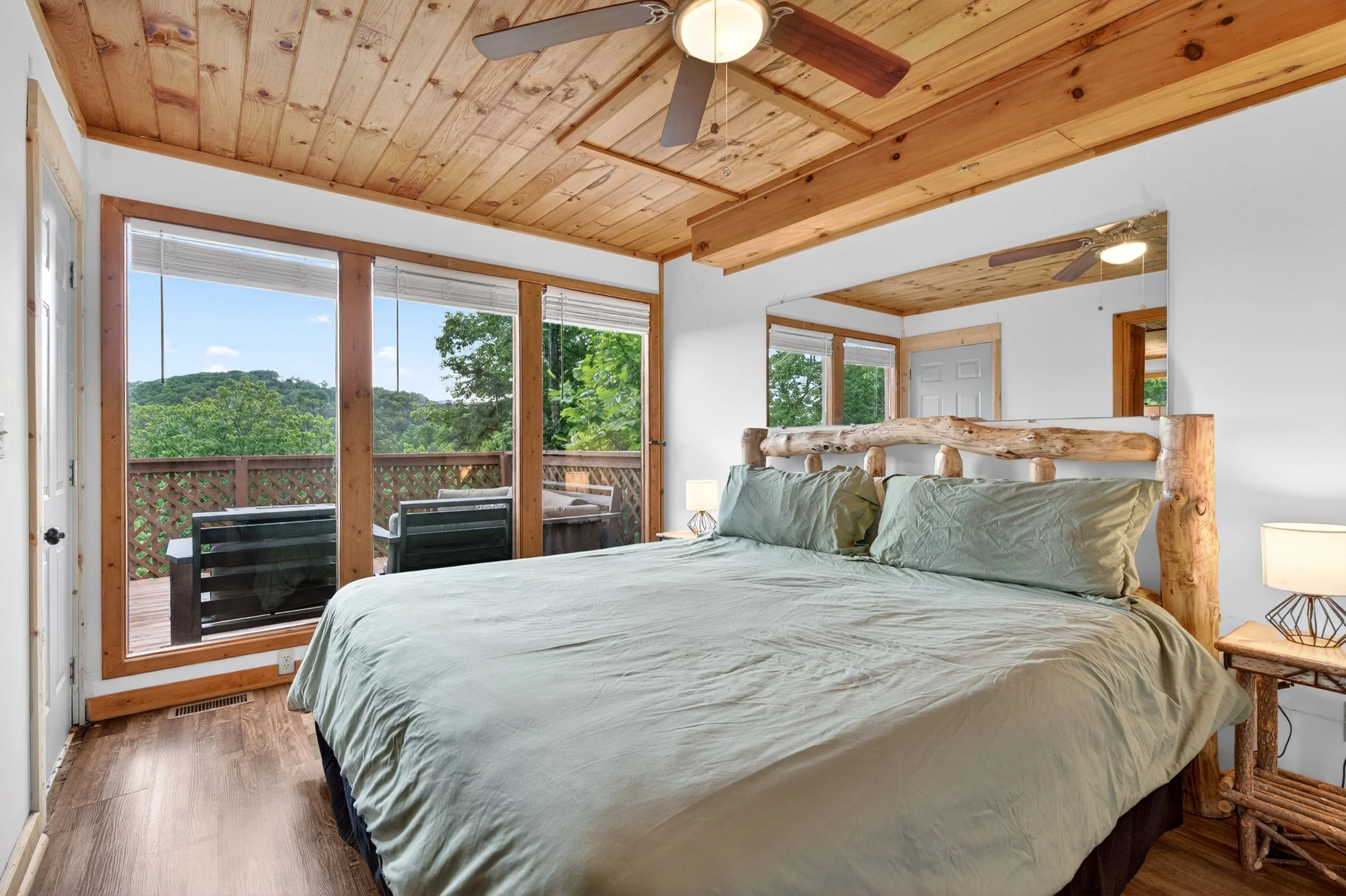 A bedroom with a rustic log bed and a wooden ceiling, large windows with a view of trees, and a wooden deck outside with patio furniture.