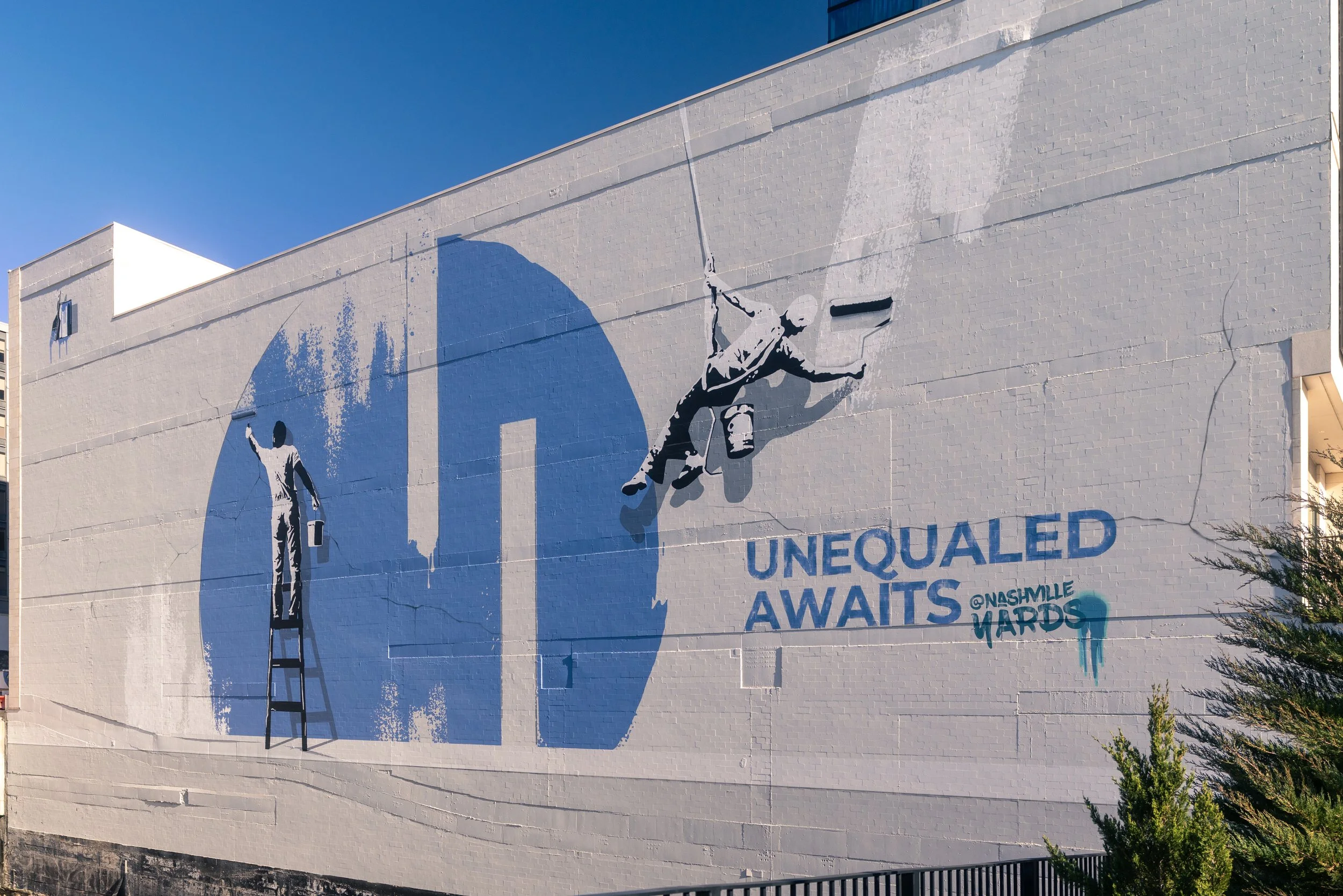 A large mural on a white brick building depicts two people painting a wall, with a ladder leaning against the mural. The mural includes the words 'INEQUAL' and 'AWAIS' with some parts obscured, and mentions '@NASHVILLE MADS.' The sky is clear and blu