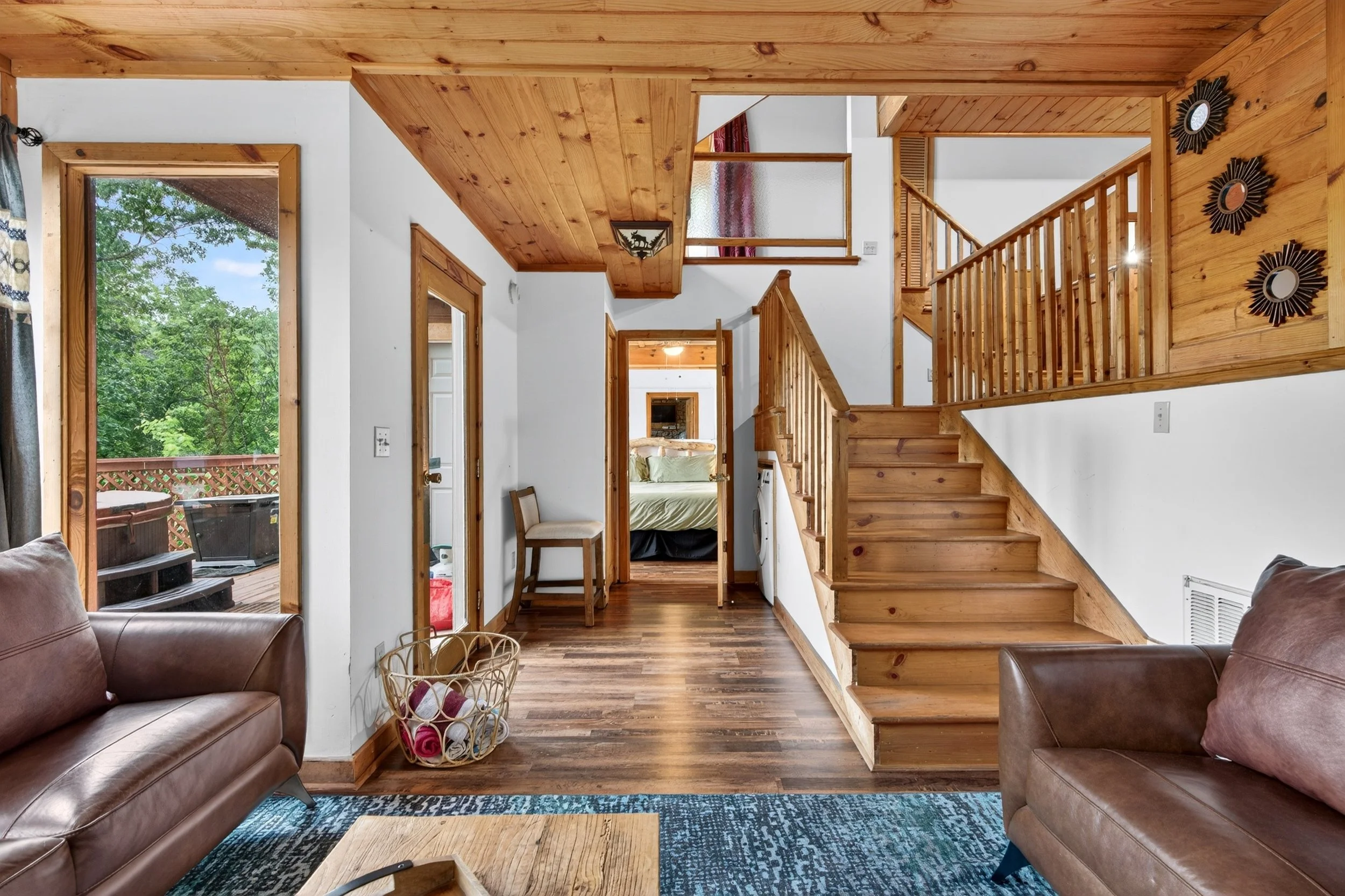 Living room with two brown leather sofas, a wooden coffee table, and a window showing outdoor trees. There is a staircase with a wooden railing leading to the upper floor, and a door to a deck outside. The ceiling and part of the wall are wooden, and