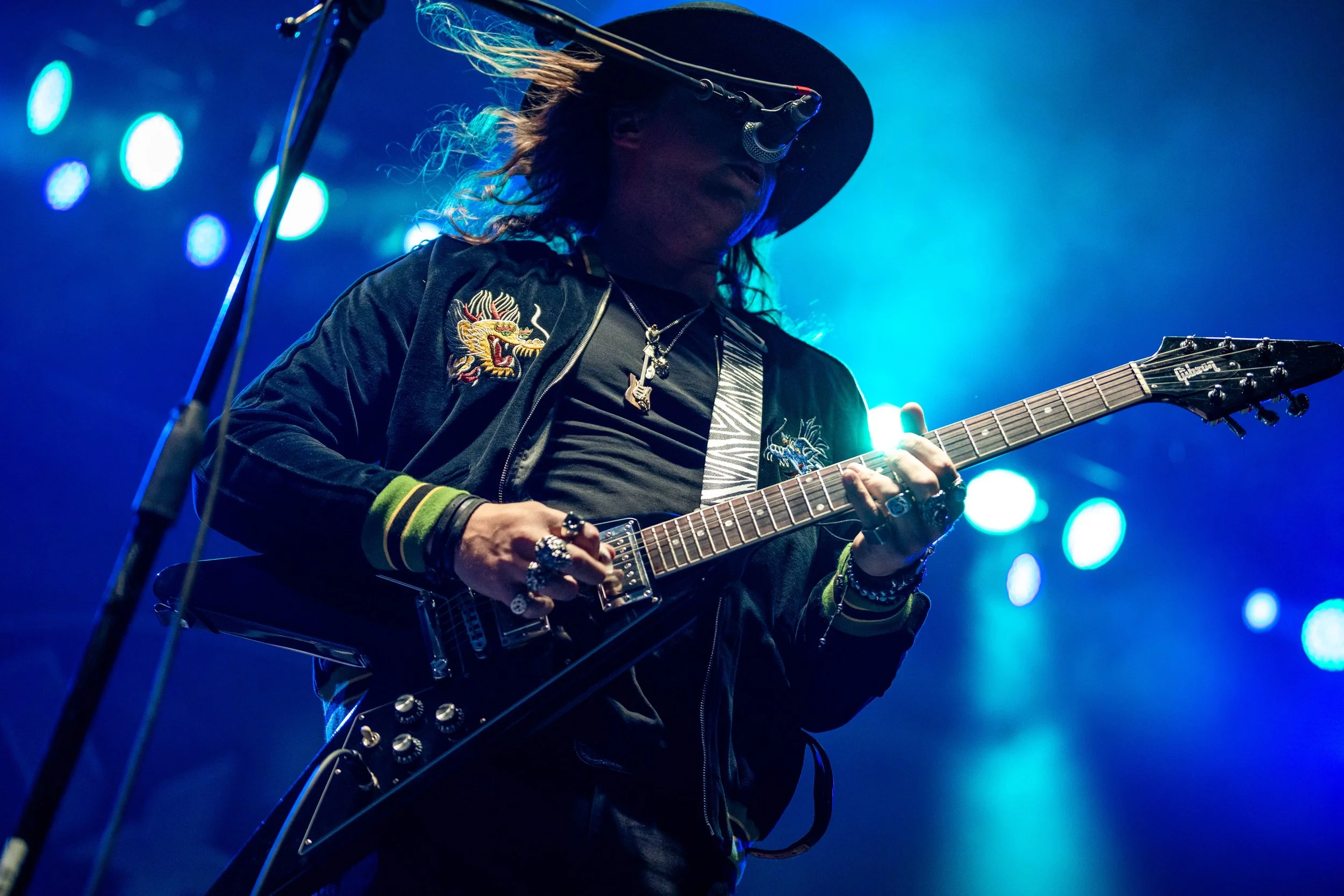 A musician performing on stage, wearing a wide-brimmed hat and dark glasses, playing an electric guitar with a black body, in front of colorful stage lights.