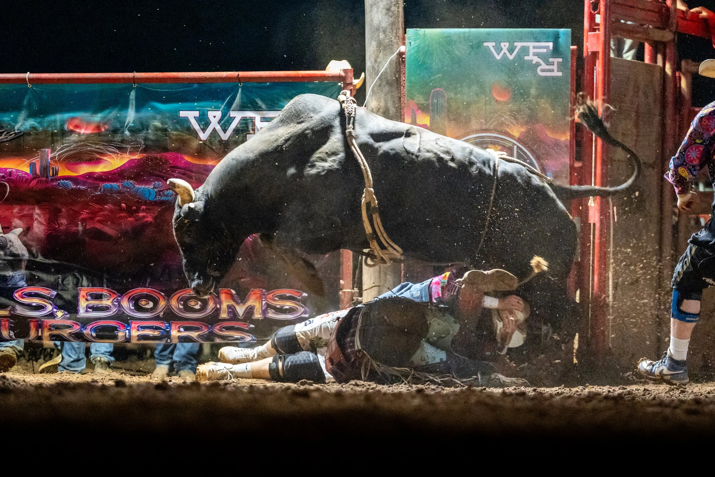 A bull rider falls off a bucking bull during a rodeo, with a woman standing close to the action, and a colorful banner with the word 'BULLS' visible in the background.
