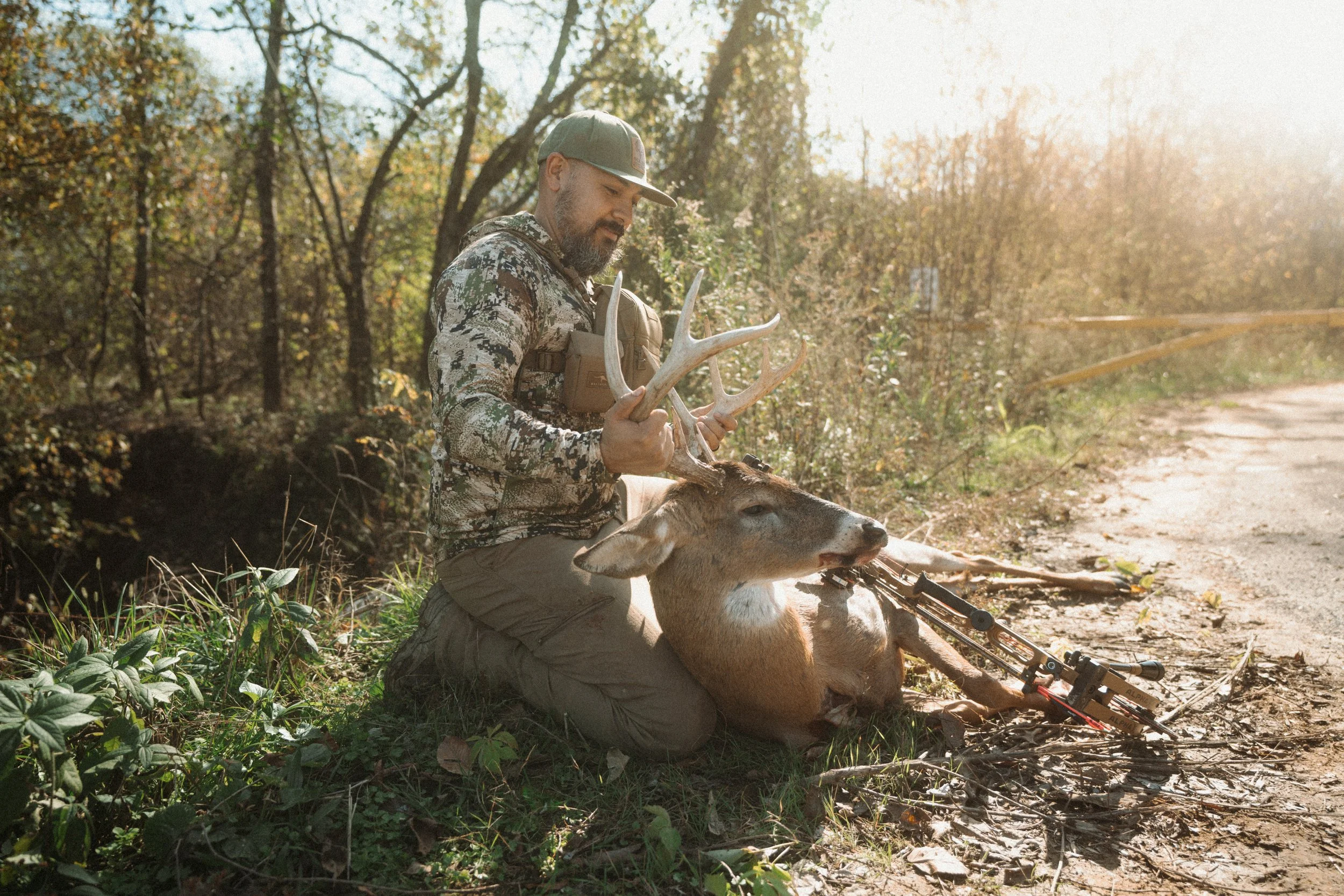 A man in camouflage clothing kneels beside a fallen deer on a dirt path in a wooded area, holding its antlers with a rifle resting on its side, during sunset or sunrise.