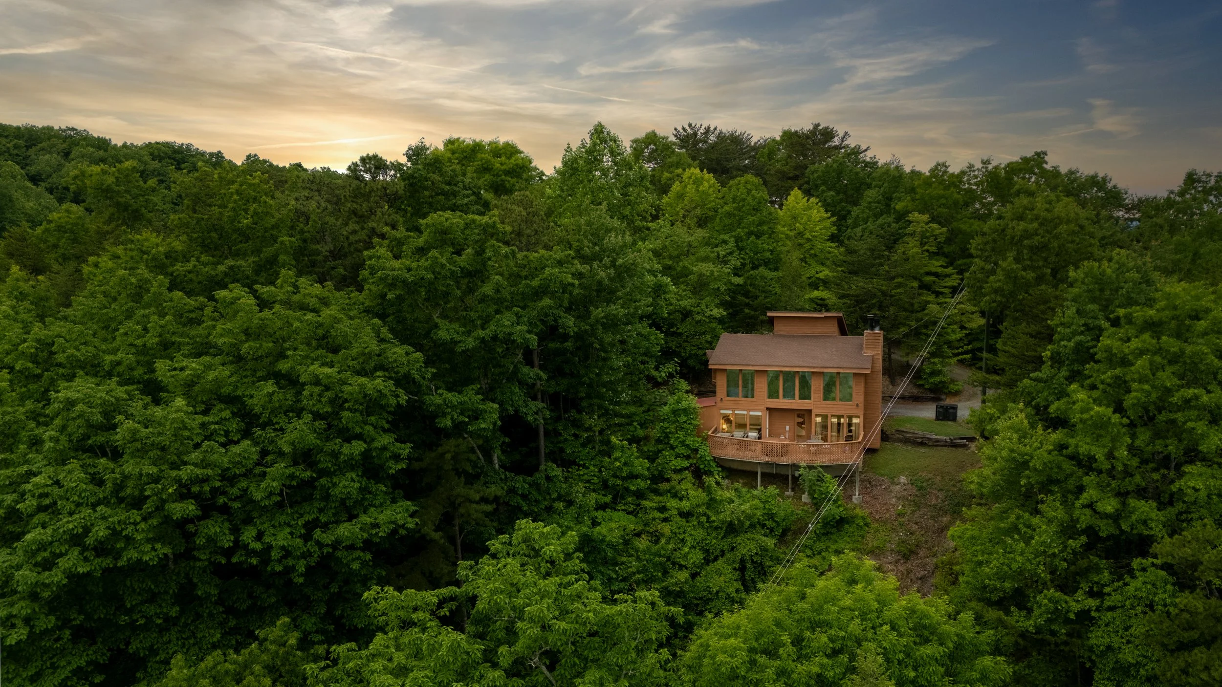 A house surrounded by dense green trees with a sunset sky in the background.