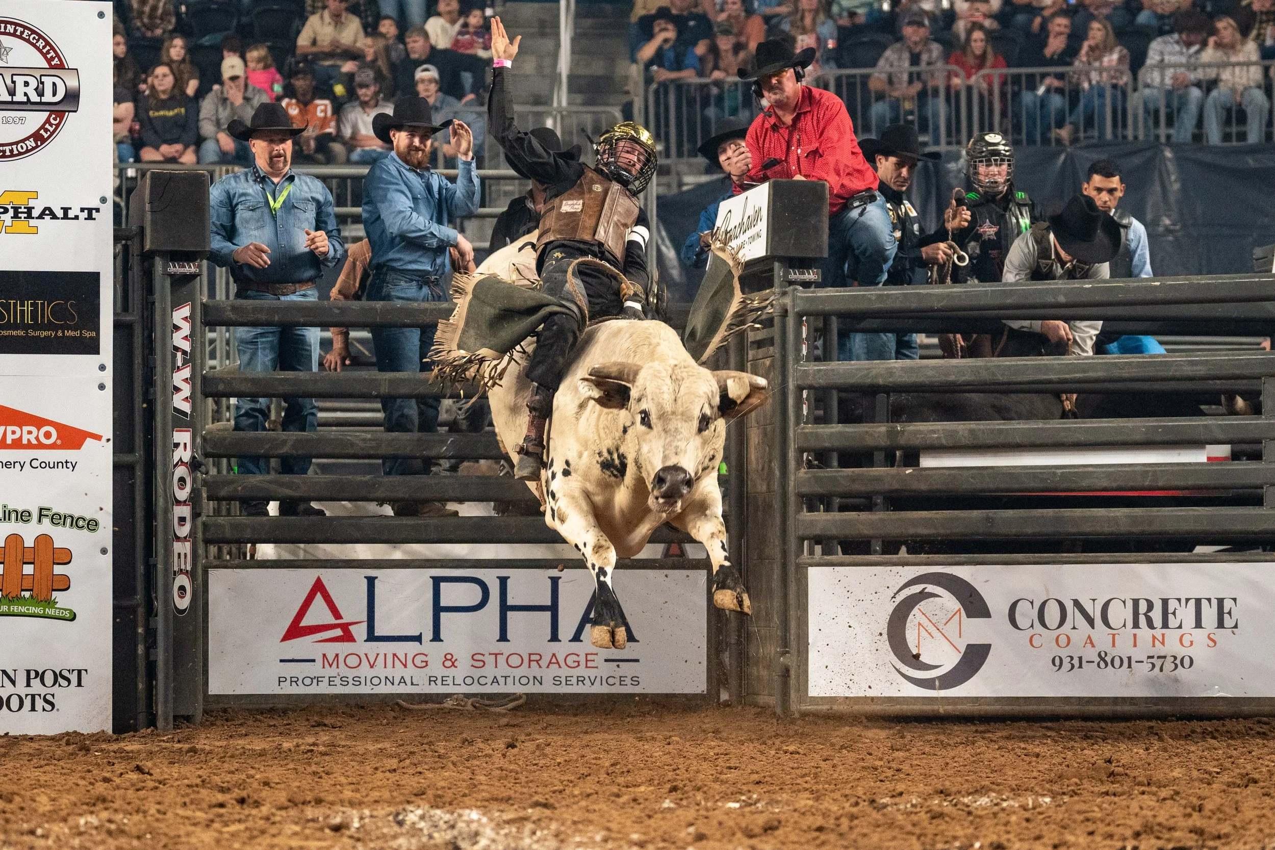 A rodeo event with a bull rider falling off a bull as it jumps over the fence, surrounded by people in western attire, with spectators in the background.