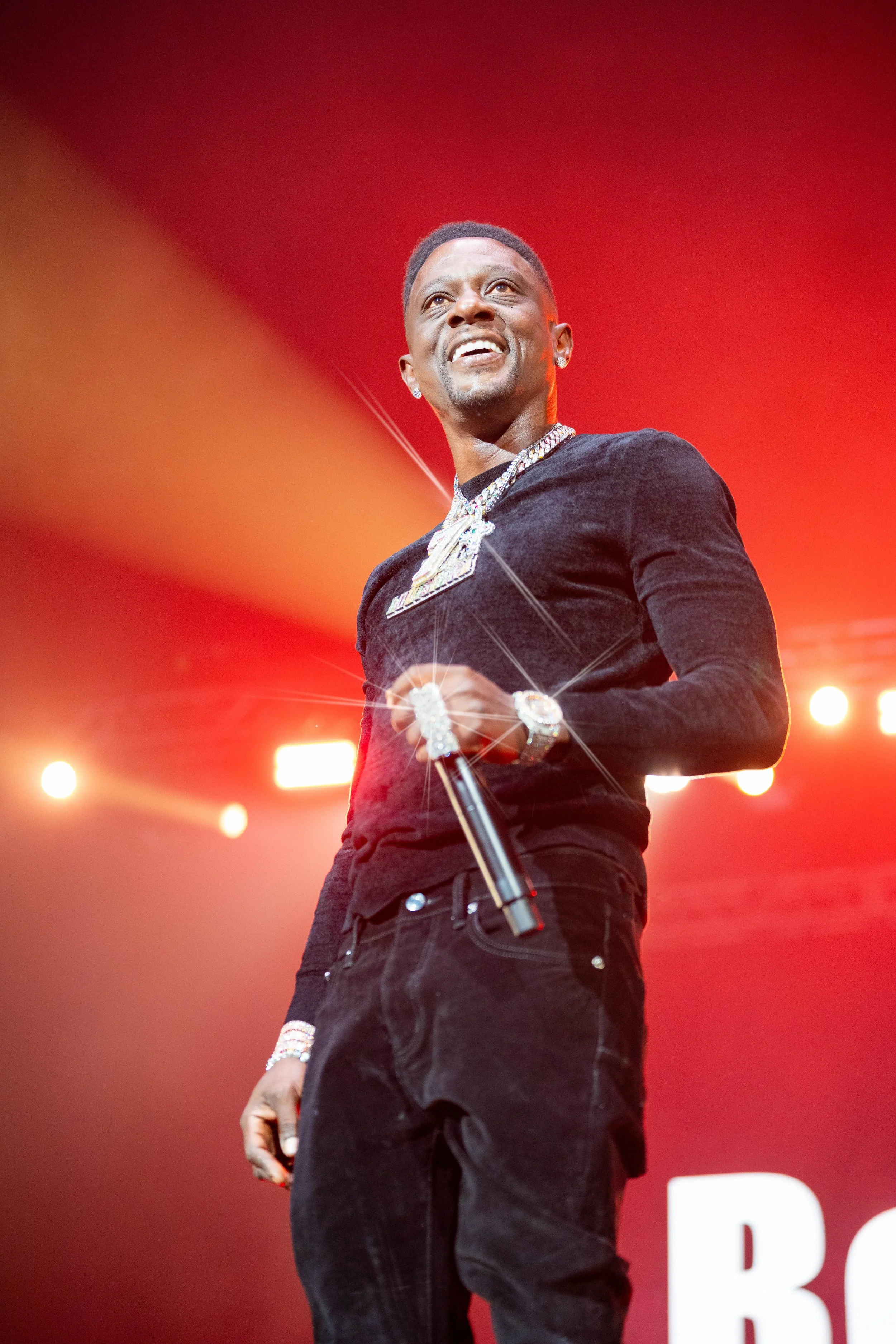 A man dressed in black with jewelry, holding a microphone on stage with bright lights and a red background.