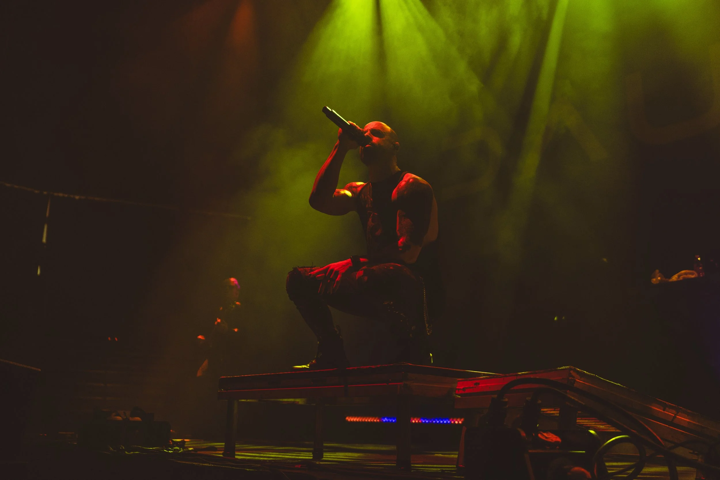 Performer kneeling on stage, singing into microphone with yellow and green stage lights overhead.