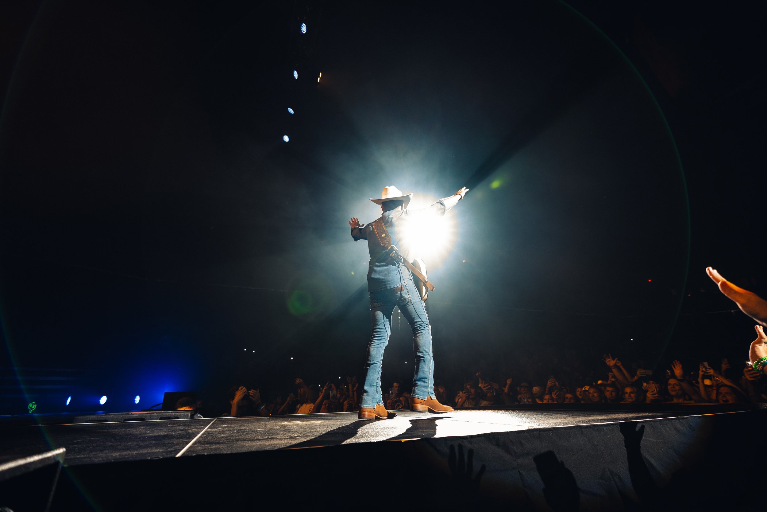 Performer wearing cowboy hat and jeans dancing on stage with bright light behind, audience reaching out hands in front.