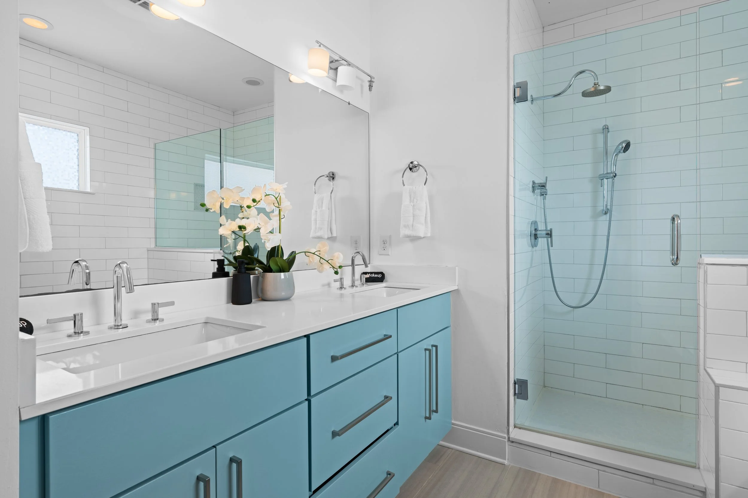 Modern bathroom with white subway tile walls, a large mirror above a white vanity with blue cabinet drawers, a flower arrangement, and a glass-enclosed shower with a rain showerhead and handheld shower attachment.