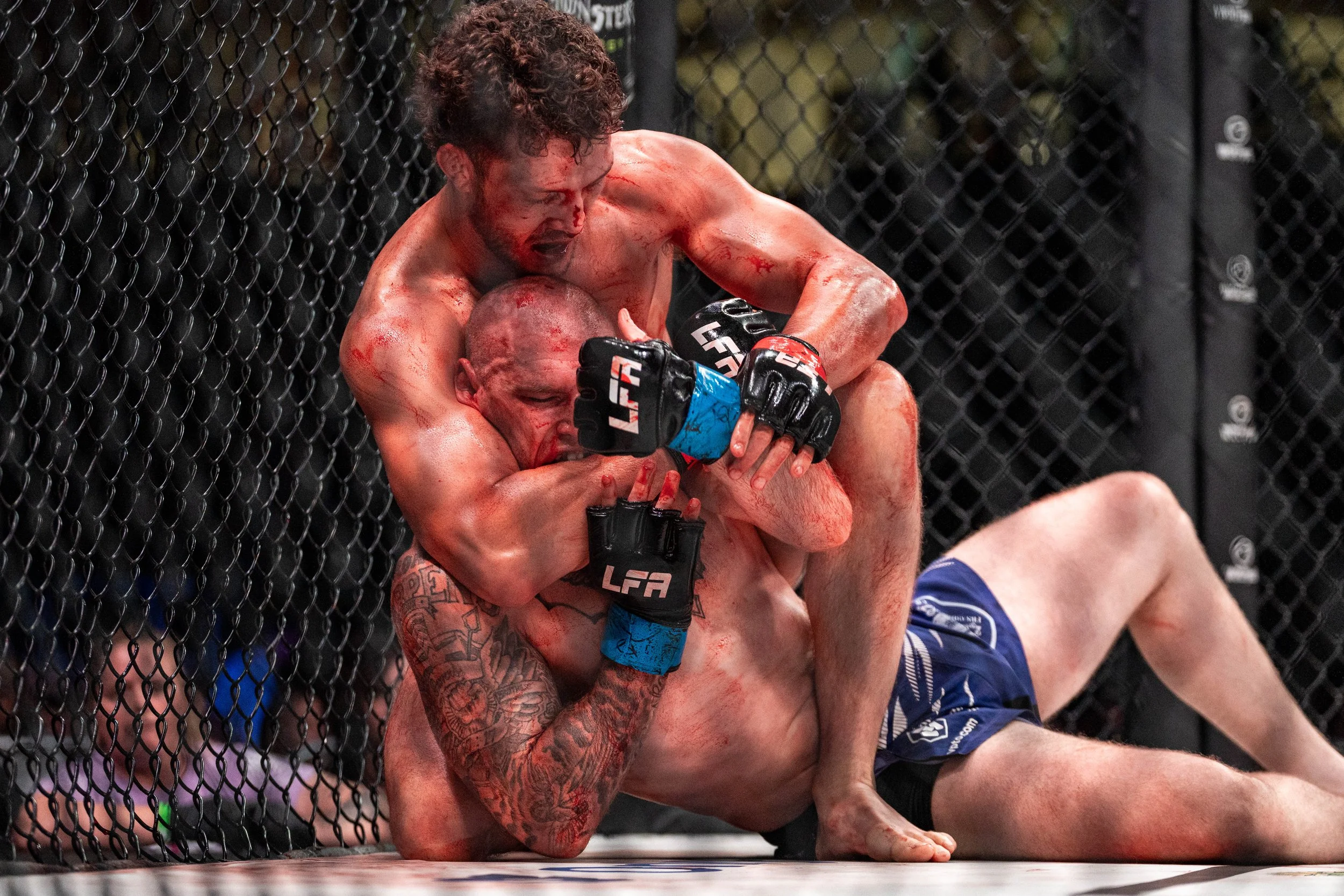 Mixed martial arts fighters grappling inside the octagon, with one fighter in black gloves applying a submission hold on the other fighter in blue gloves. The fighters are against the cage, and blood is visible on both fighters.