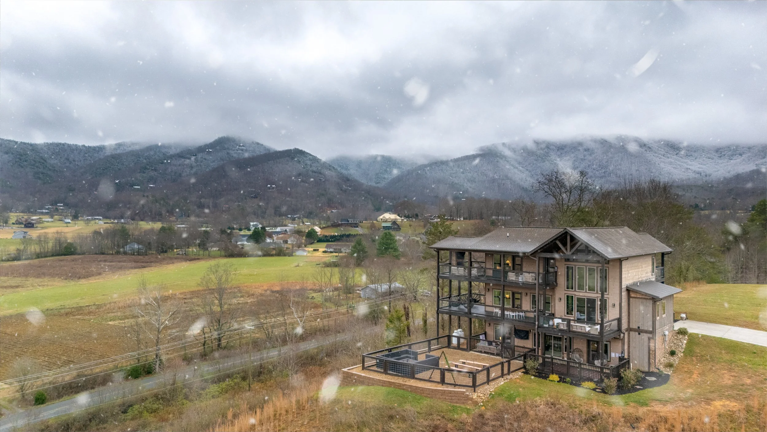 View of a mountainous landscape on a rainy day, with a modern multi-story house in the foreground and snow falling from a cloudy sky.
