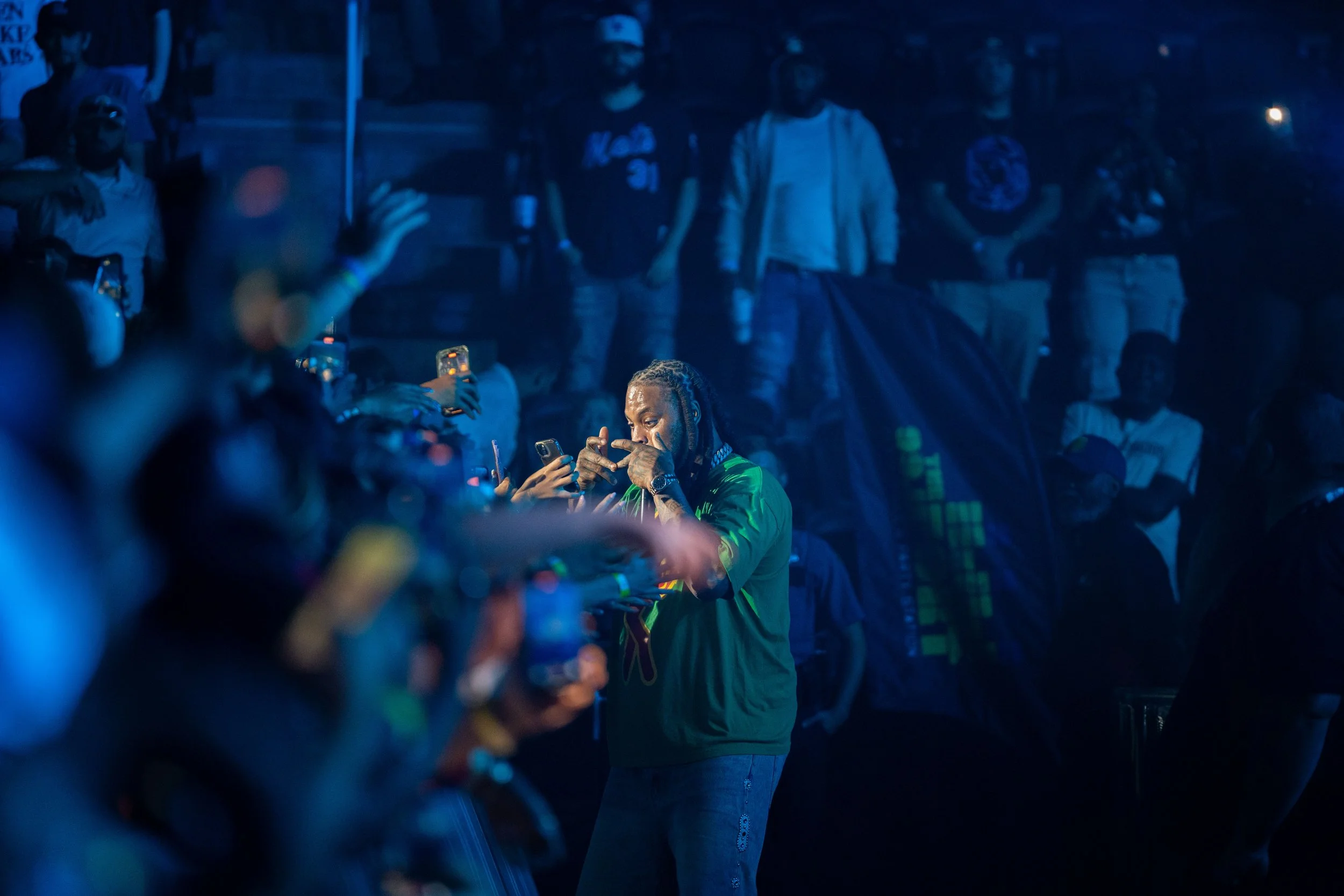 A man with braided hair and a green jacket is taking a photo of himself with his phone at a nighttime outdoor event, surrounded by people, some holding their phones, with a dark background and blue lighting.