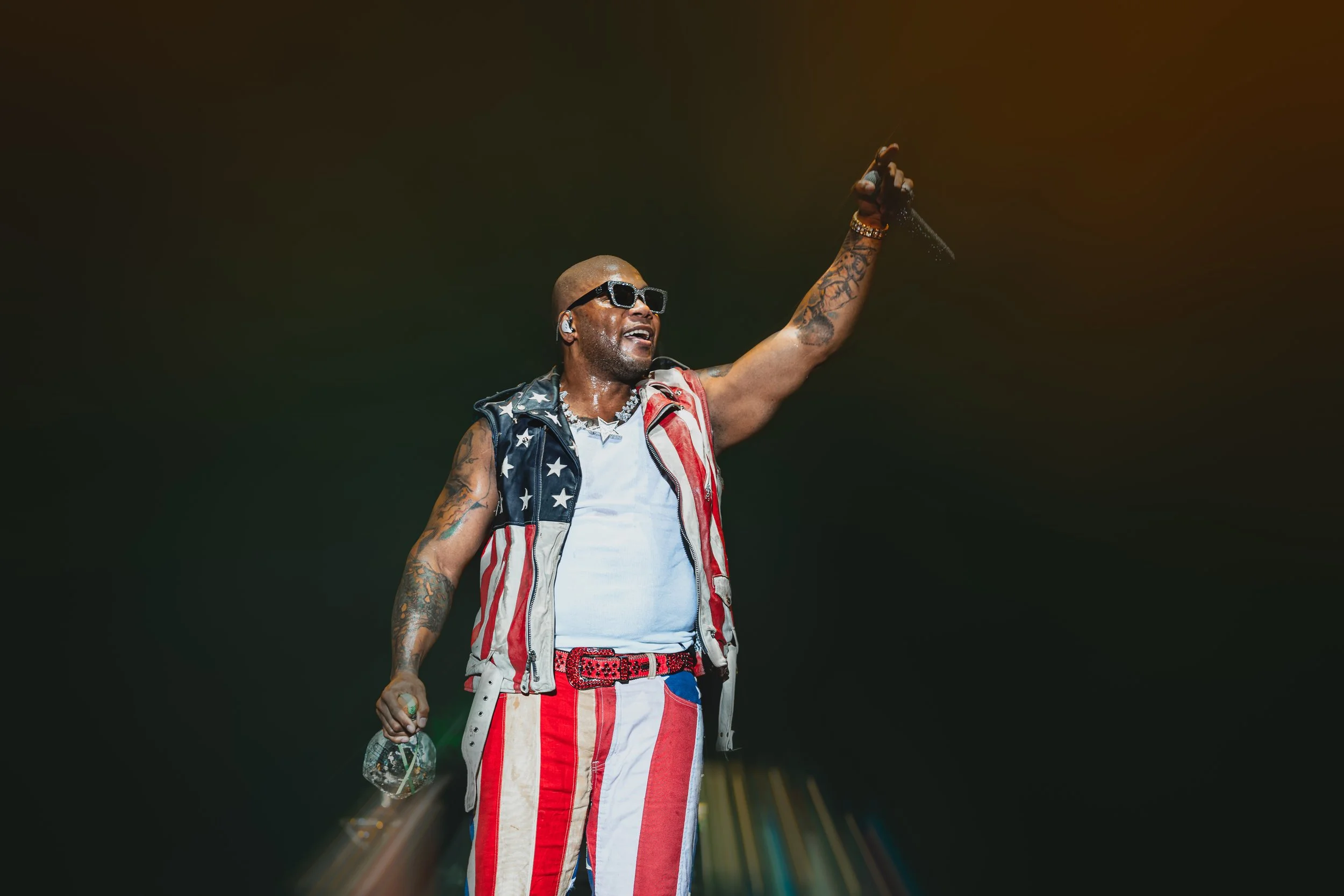 A male performer on stage, wearing sunglasses, a sleeveless vest with an American flag design, and red, white, and blue striped pants. He is bald, has tattoos on his arms, and is holding a microphone in his left hand while raising his right hand.