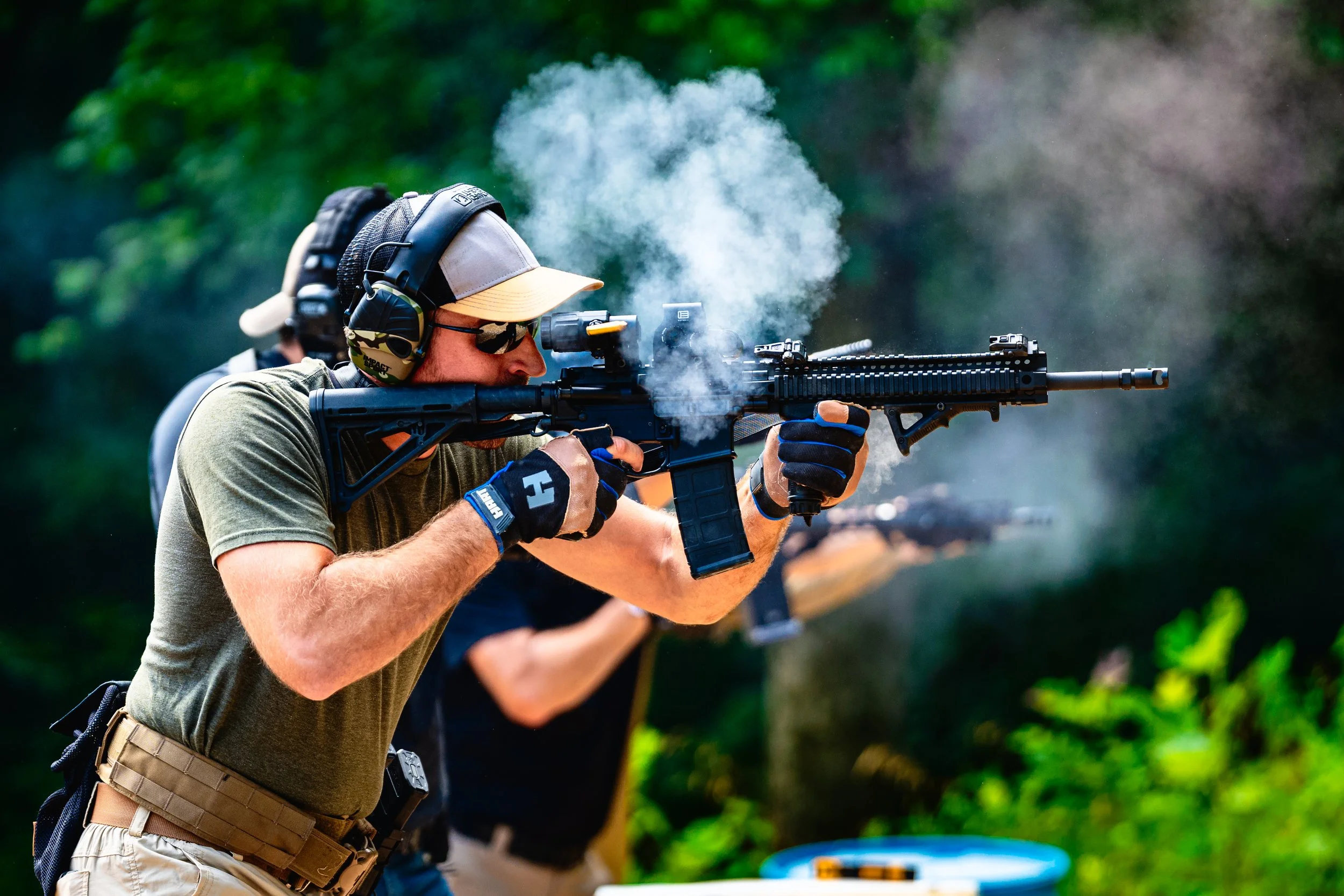 Man wearing sunglasses, a cap, and hearing protection, aiming a rifle during a shooting event outdoors with green trees in the background.