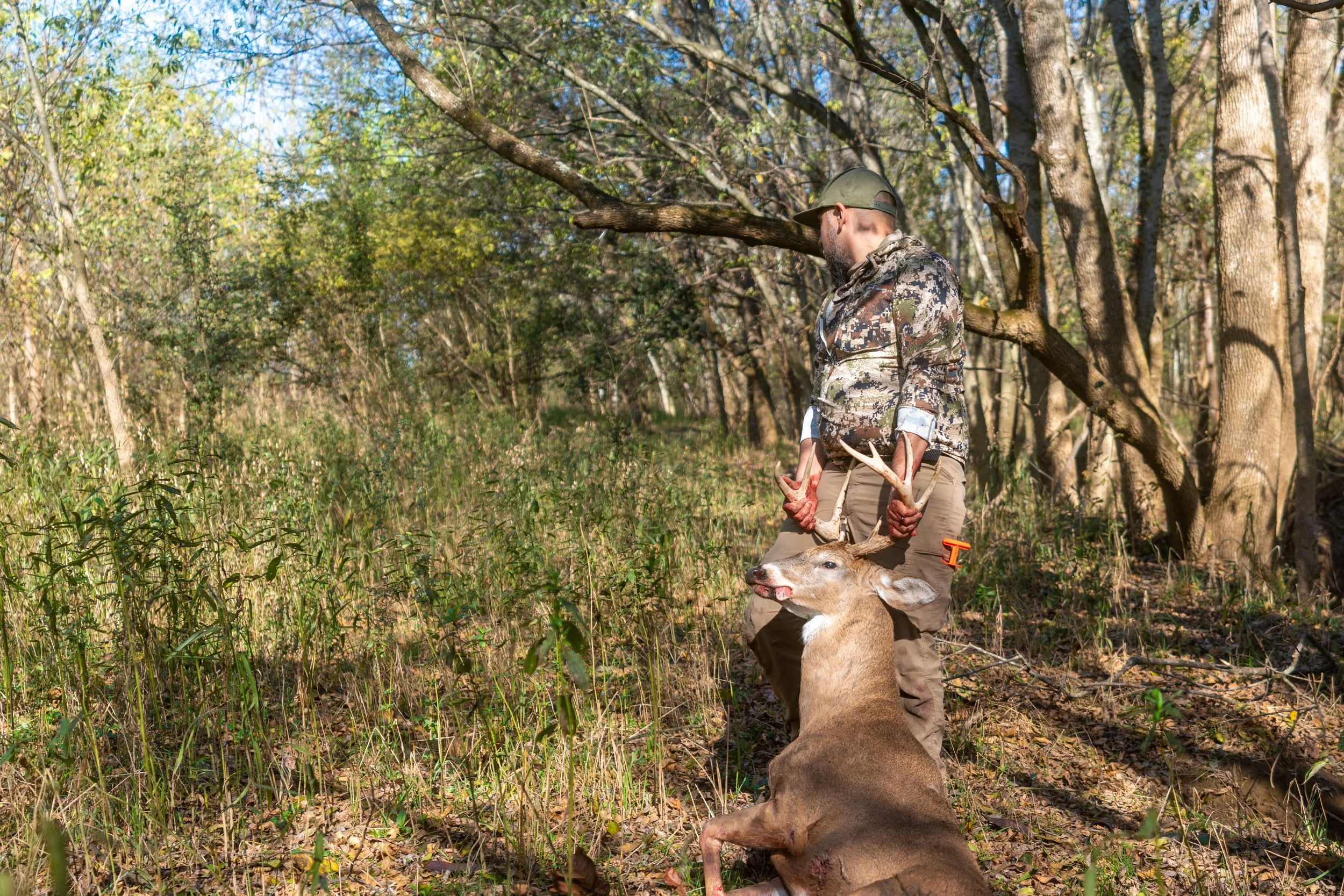 Man in camouflage clothing holding a deer by its antlers in a forested area with trees and green foliage.
